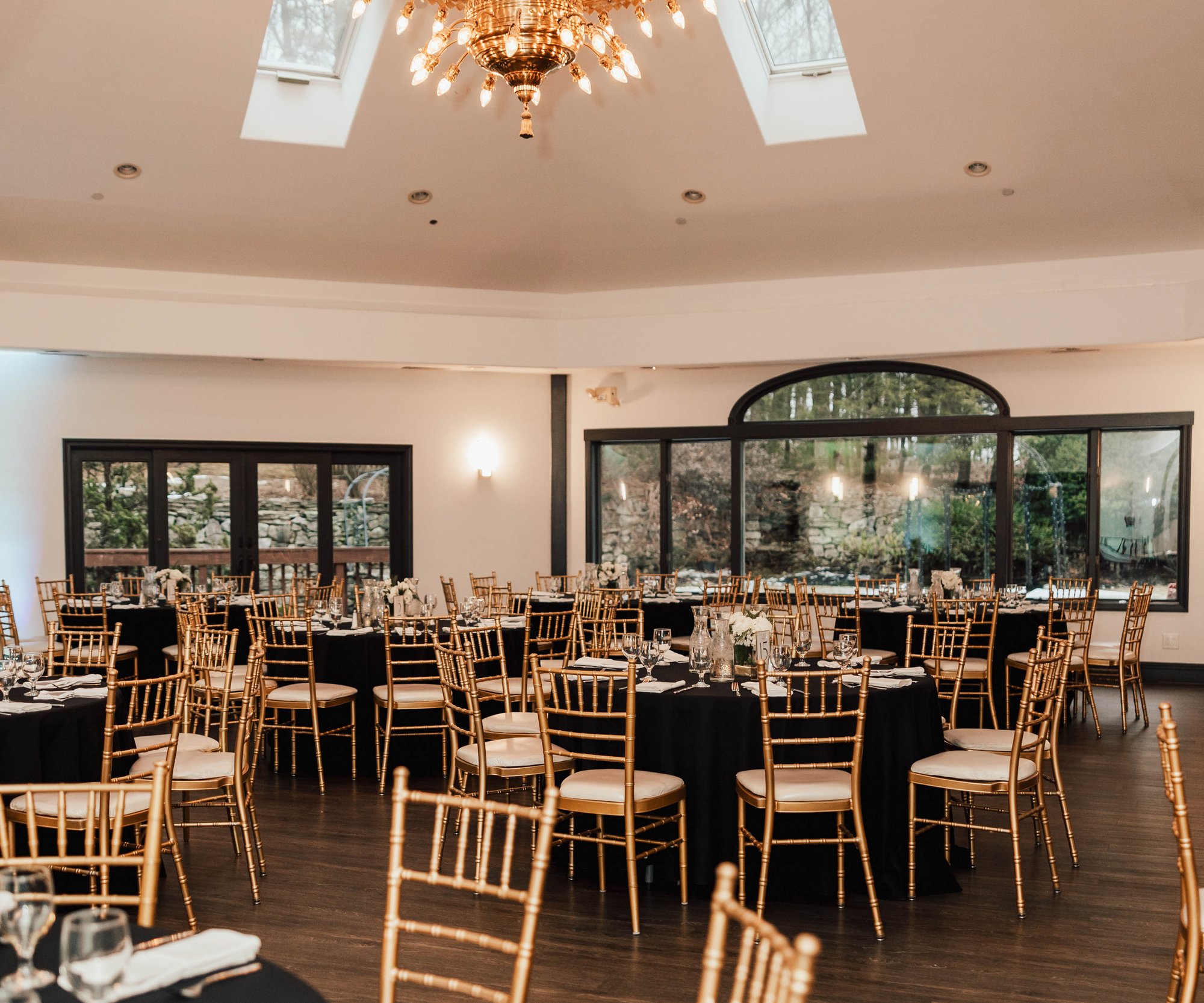 An elegant reception hall at Granite Rose set with round tables covered in black tablecloths and gold chiavari chairs. Large windows showcase the surrounding woodland. The space features white walls, wooden flooring, and is illuminated by a grand gold chandelier with skylights above.