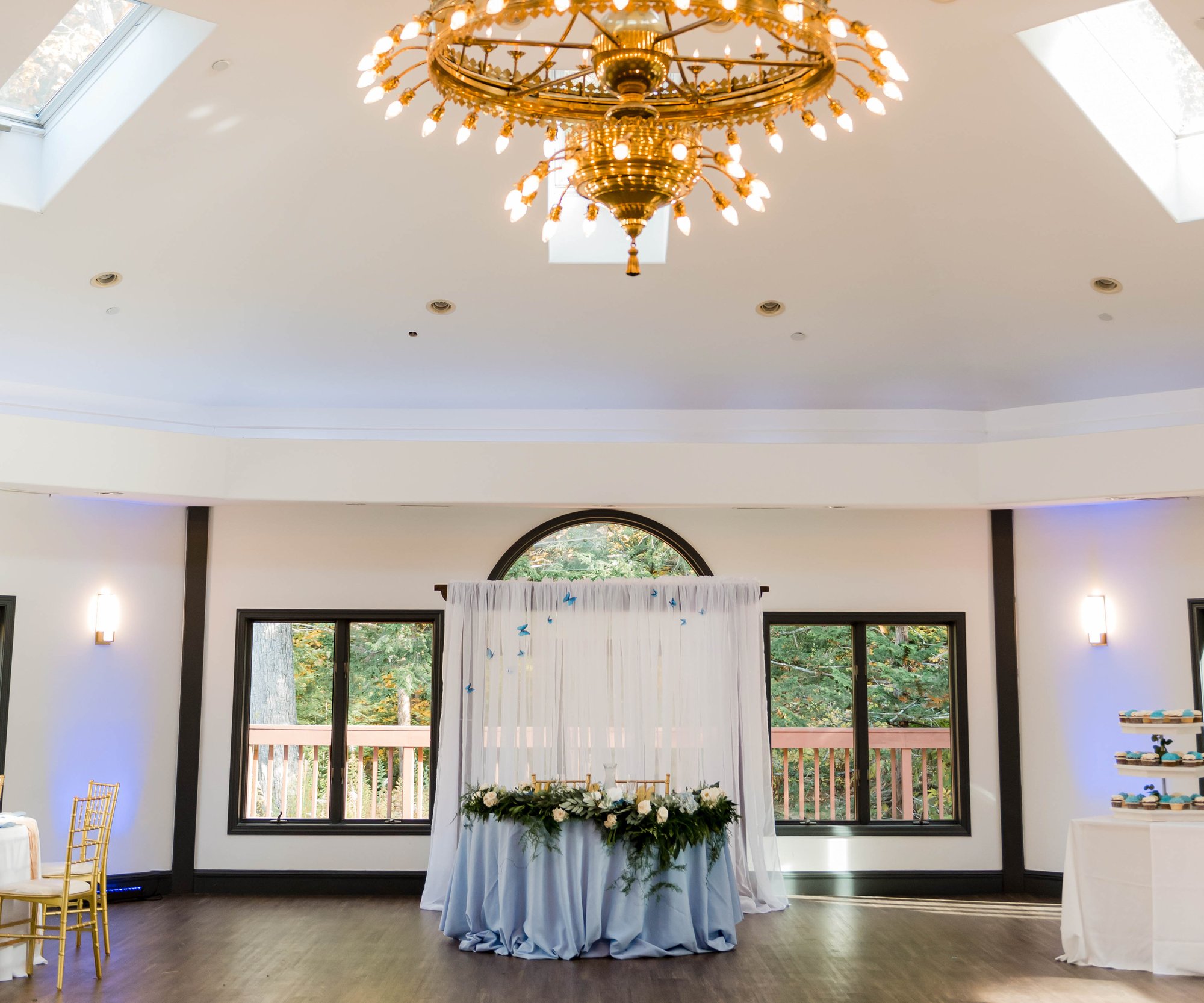 An elegant wedding sweetheart table at Granite Rose featuring a round table with floral arrangements under a grand gold chandelier. White draping creates a backdrop between two windows with forest views. The room has white walls, wooden flooring, and a high ceiling with skylights.