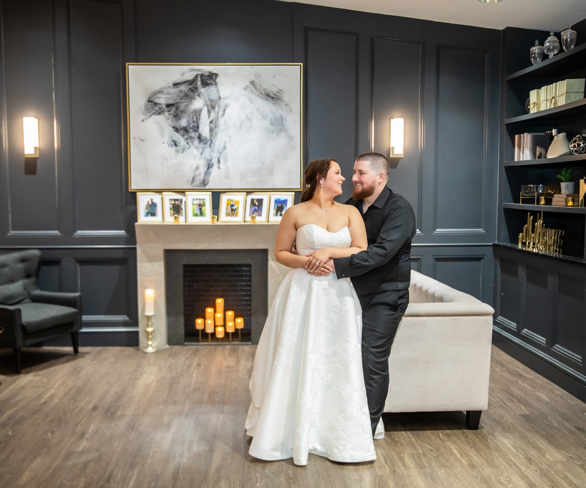 A cozy lounge area at Granite Rose featuring dark navy paneled walls, a fireplace with candles, modern abstract artwork, and comfortable seating. The wooden flooring complements the elegant decor with gold accents, built-in shelving, and warm lighting from wall sconces.