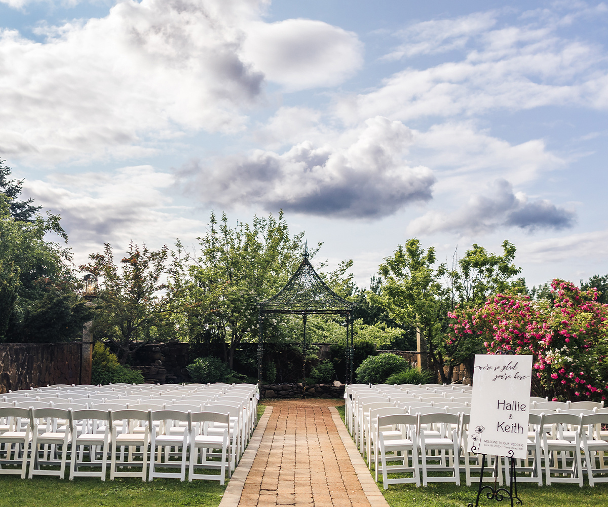 An outdoor wedding ceremony setup at Granite Rose by Wedgewood Weddings. Two sections of white folding chairs create an aisle leading to a black metal gazebo. The gazebo is surrounded by lush green trees and landscaped gardens with some pink flowering bushes visible on the right side.