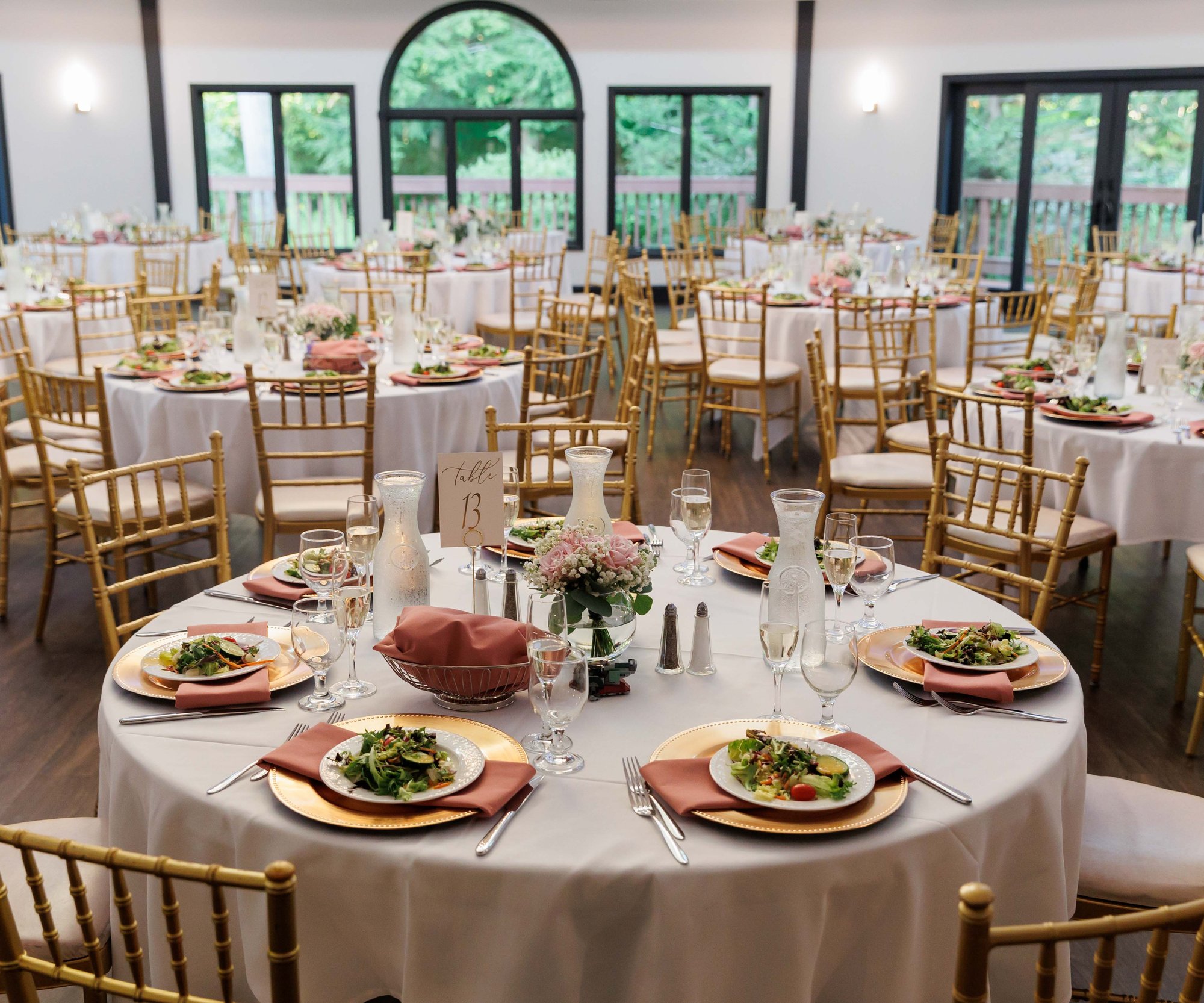 A beautifully set wedding reception hall with round tables covered in white tablecloths, gold chiavari chairs, and coral/pink napkins. Each table features salad plates, water glasses, wine glasses, and small floral centerpieces. Large windows reveal a garden view.