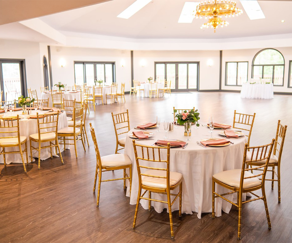 An elegant indoor reception hall at Granite Rose. The spacious room features a polished wood floor and white walls with large windows letting in natural light. Each table is surrounded by gold chiavari chairs and features a small floral centerpiece with colorful flowers.