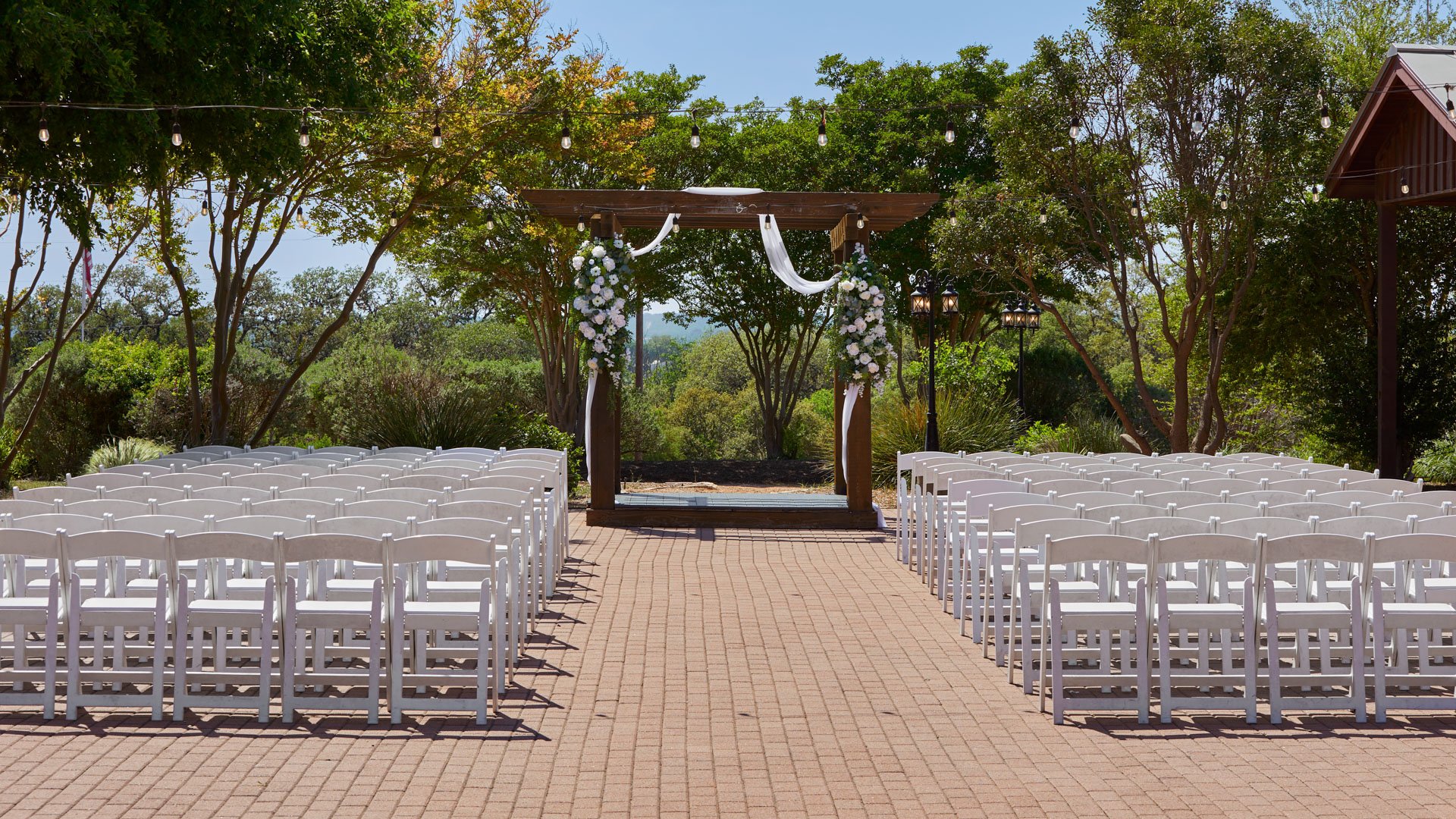 Elegant outdoor wedding ceremony setup on the brick patio at Hofmann Ranch in Castroville, TX, featuring white chairs, floral arch décor, and lush green surroundings.