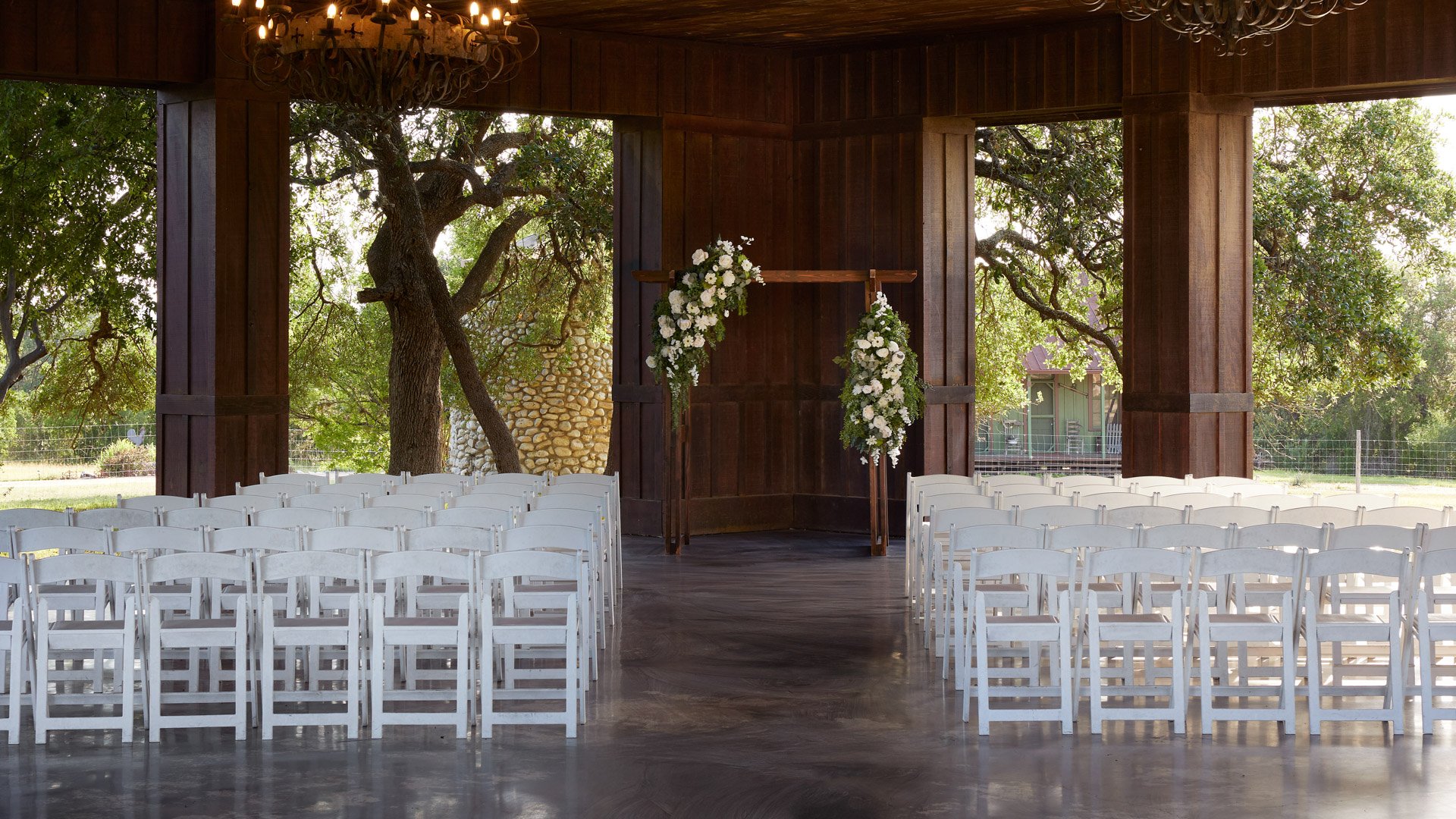Wedding ceremony inside the Meadow Pavilion at Hofmann Ranch, featuring white seating, floral arch décor, rustic chandeliers, and countryside views through open walls.