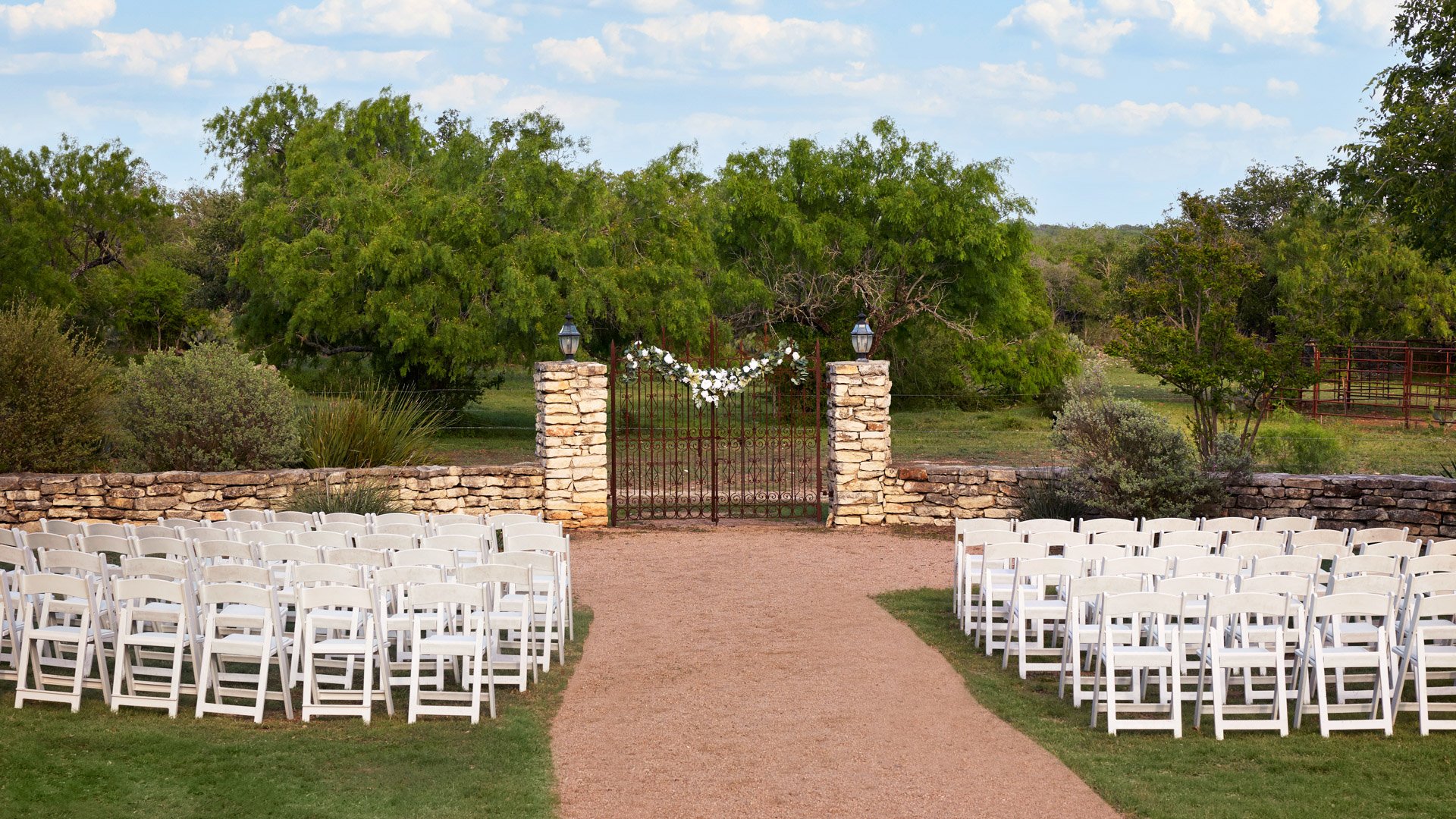 White chairs arranged for a wedding ceremony facing the iconic stone and iron gates at Hofmann Ranch in Castroville, TX.