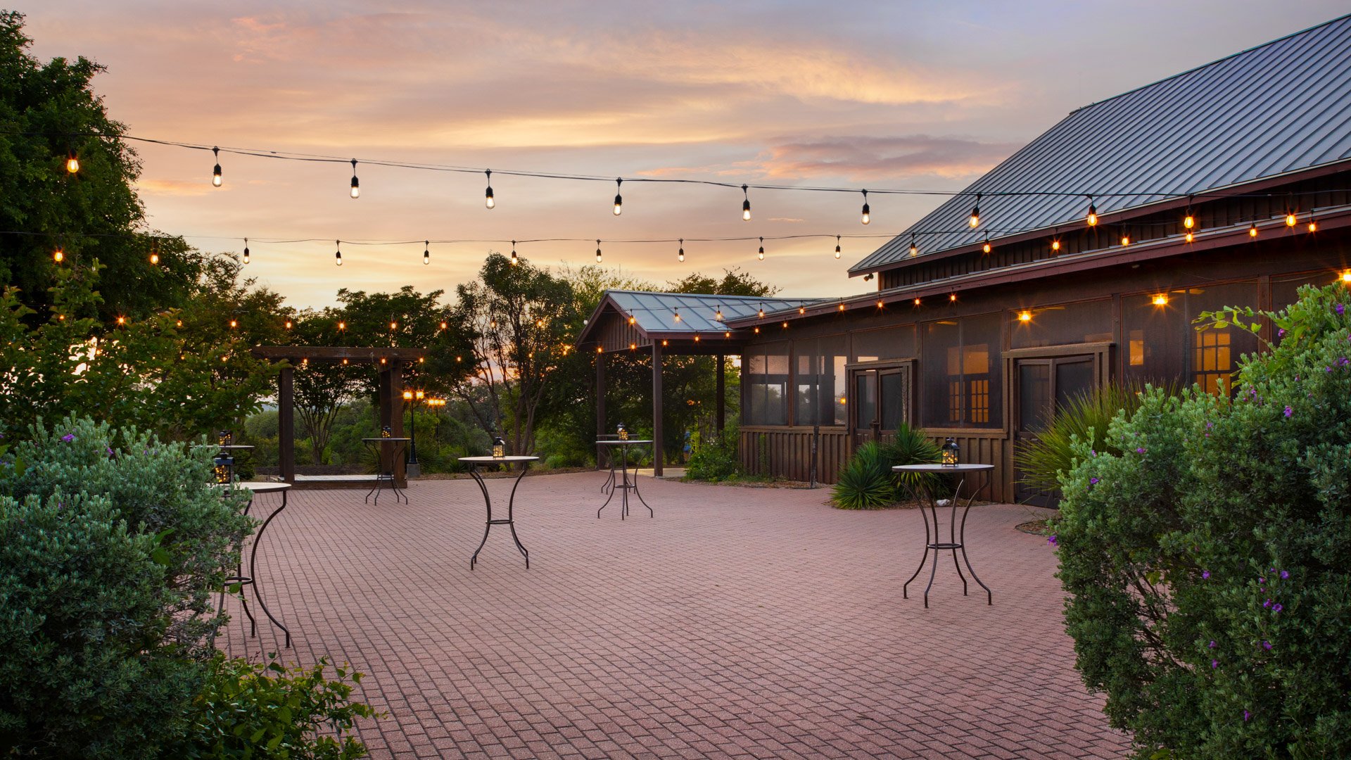 Evening cocktail hour on the brick patio at Hofmann Ranch with string lights glowing against a Texas sunset.