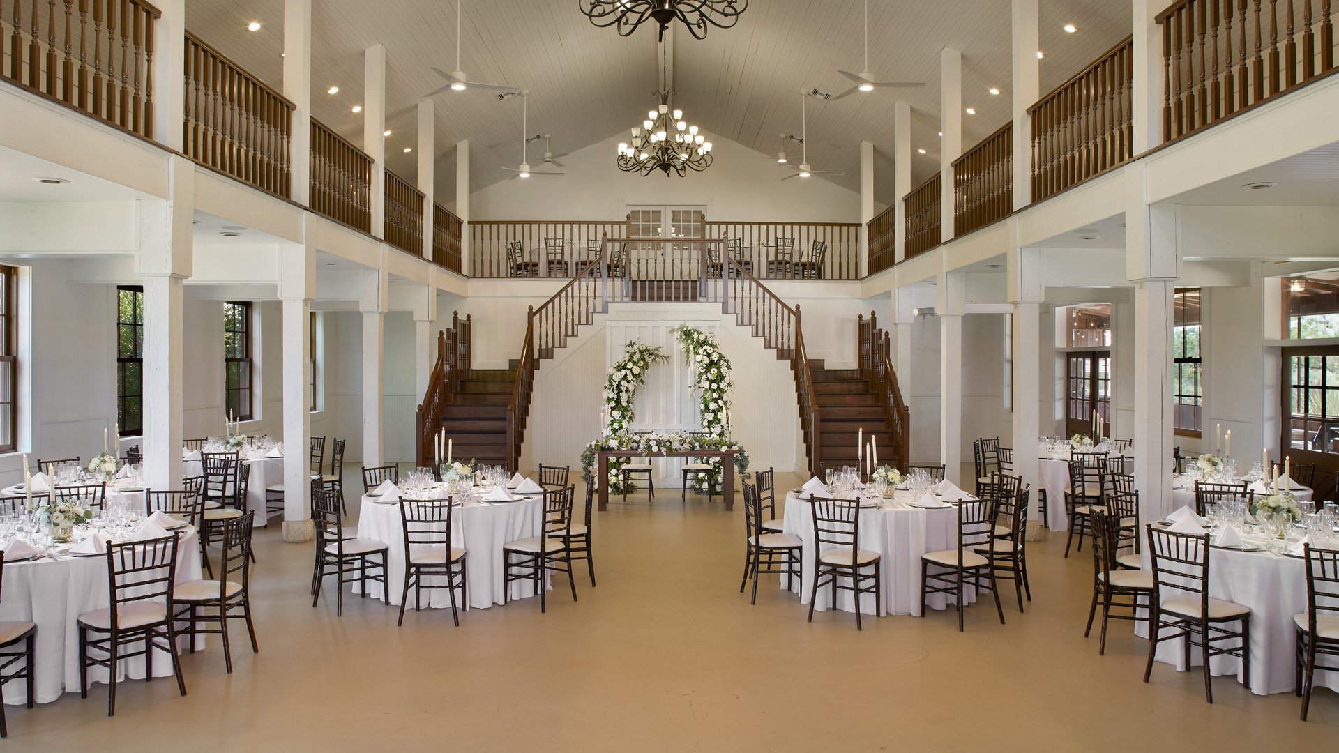 The Grande Hall at Hofmann Ranch set for a wedding reception with round tables, white linens, floral centerpieces, and dramatic dual staircases as the backdrop.