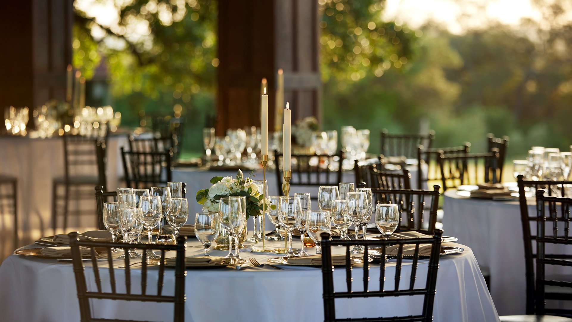 Close-up of a wedding reception table at Hofmann Ranch with white linens, glassware, candles, and floral centerpiece under warm sunlight.