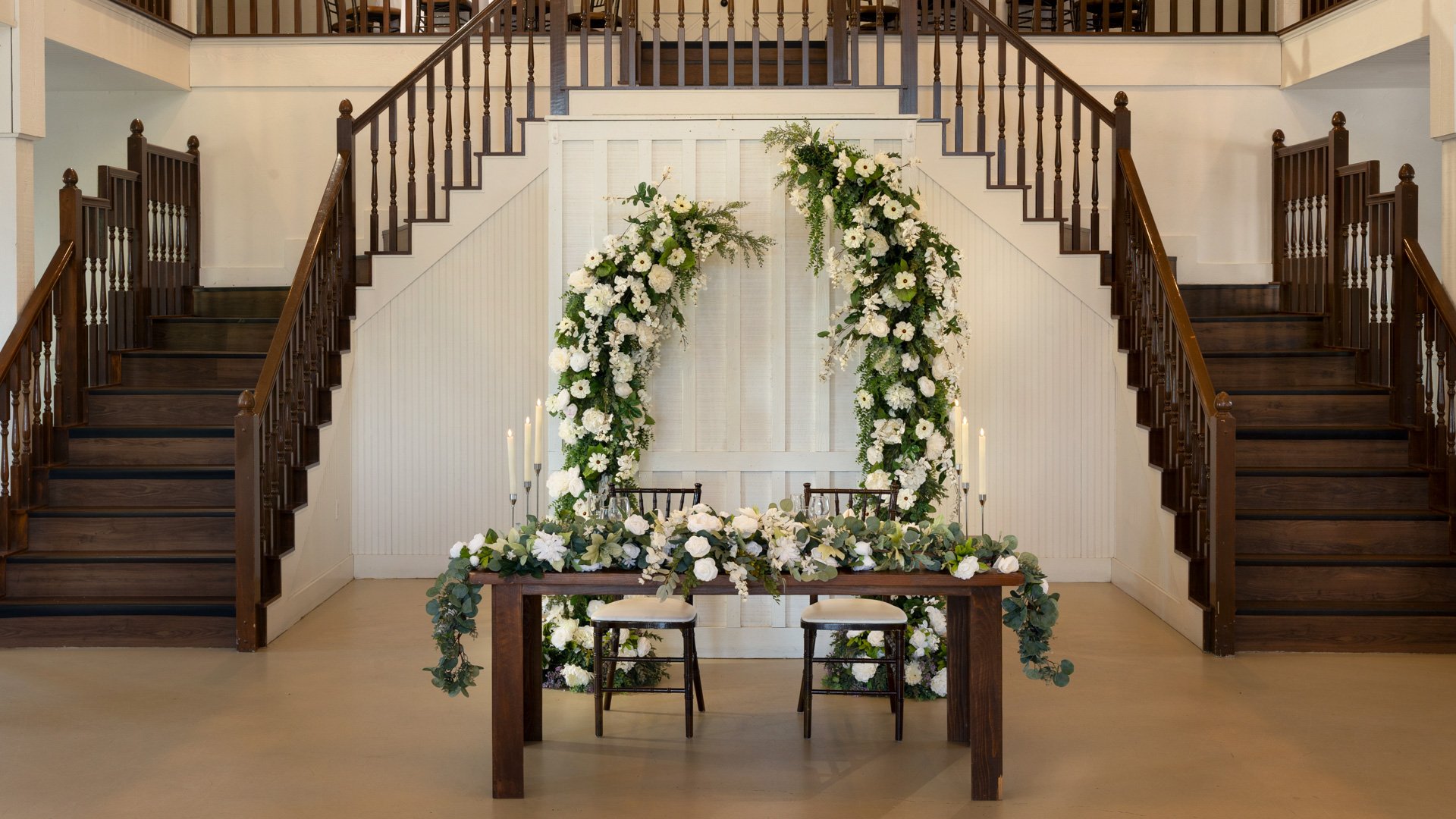 Romantic sweetheart table in the Grande Hall at Hofmann Ranch featuring white floral arch, lush greenery, and classic wooden staircase backdrop.