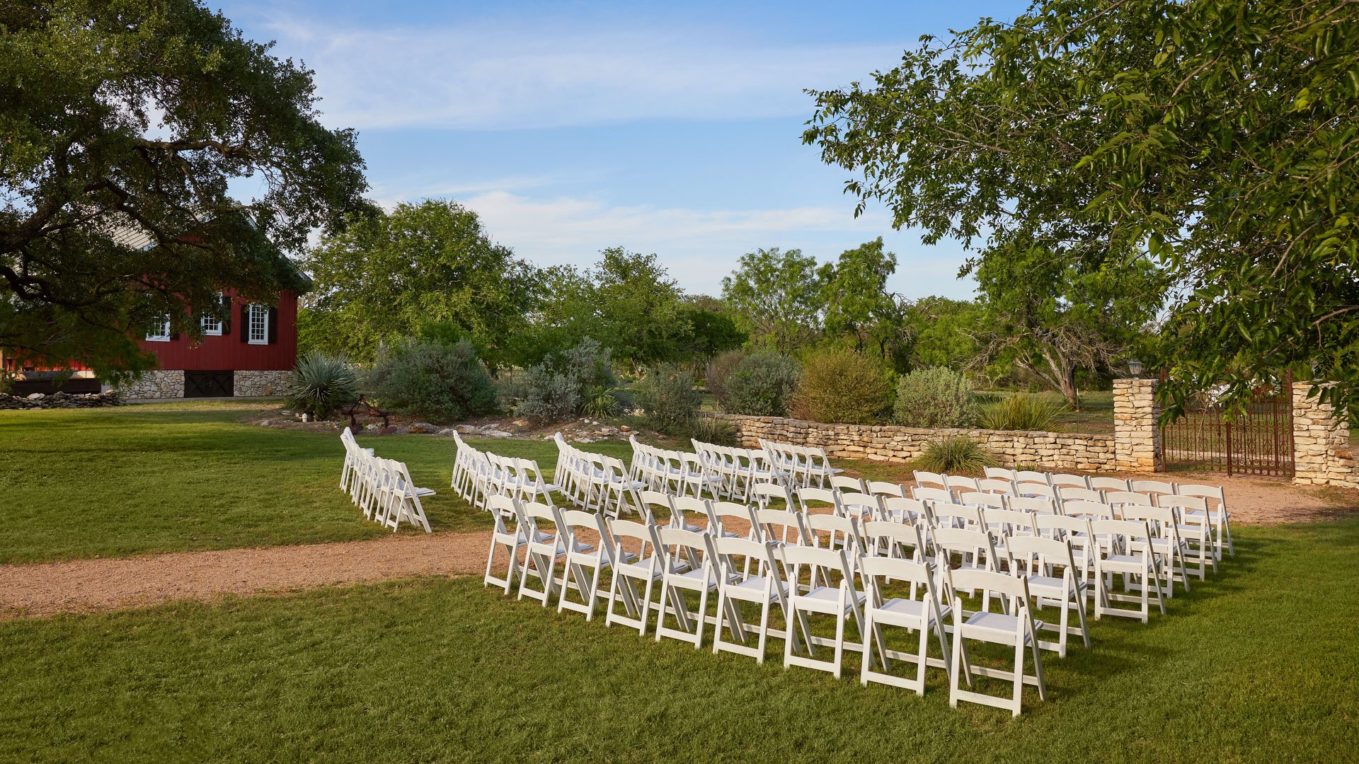 Outdoor ceremony space at Hofmann Ranch featuring white chairs, rustic iron gates, and a scenic red barn in the background.