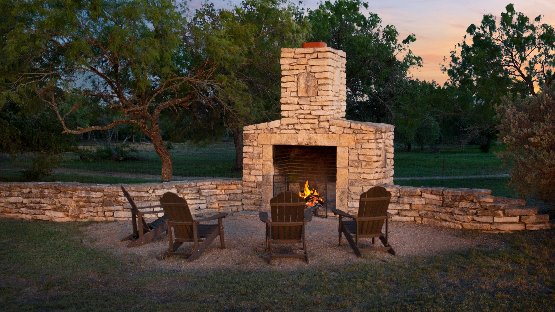 Cozy outdoor stone fireplace at Hofmann Ranch with Adirondack chairs set under the Texas evening sky.