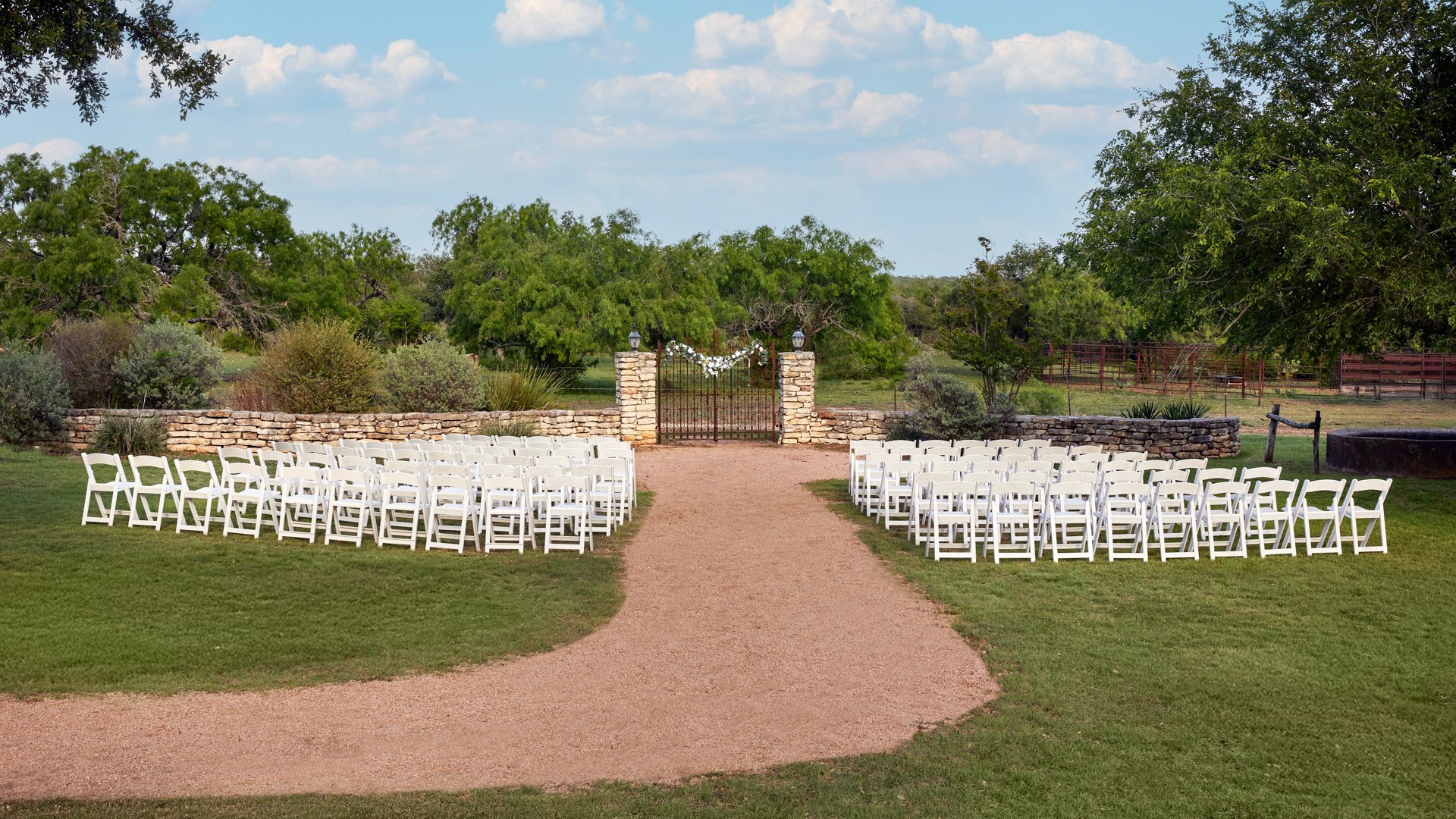 Outdoor wedding ceremony at Hofmann Ranch in Castroville, TX, with white chairs facing a rustic stone and iron gate altar framed by Texas hill country scenery.