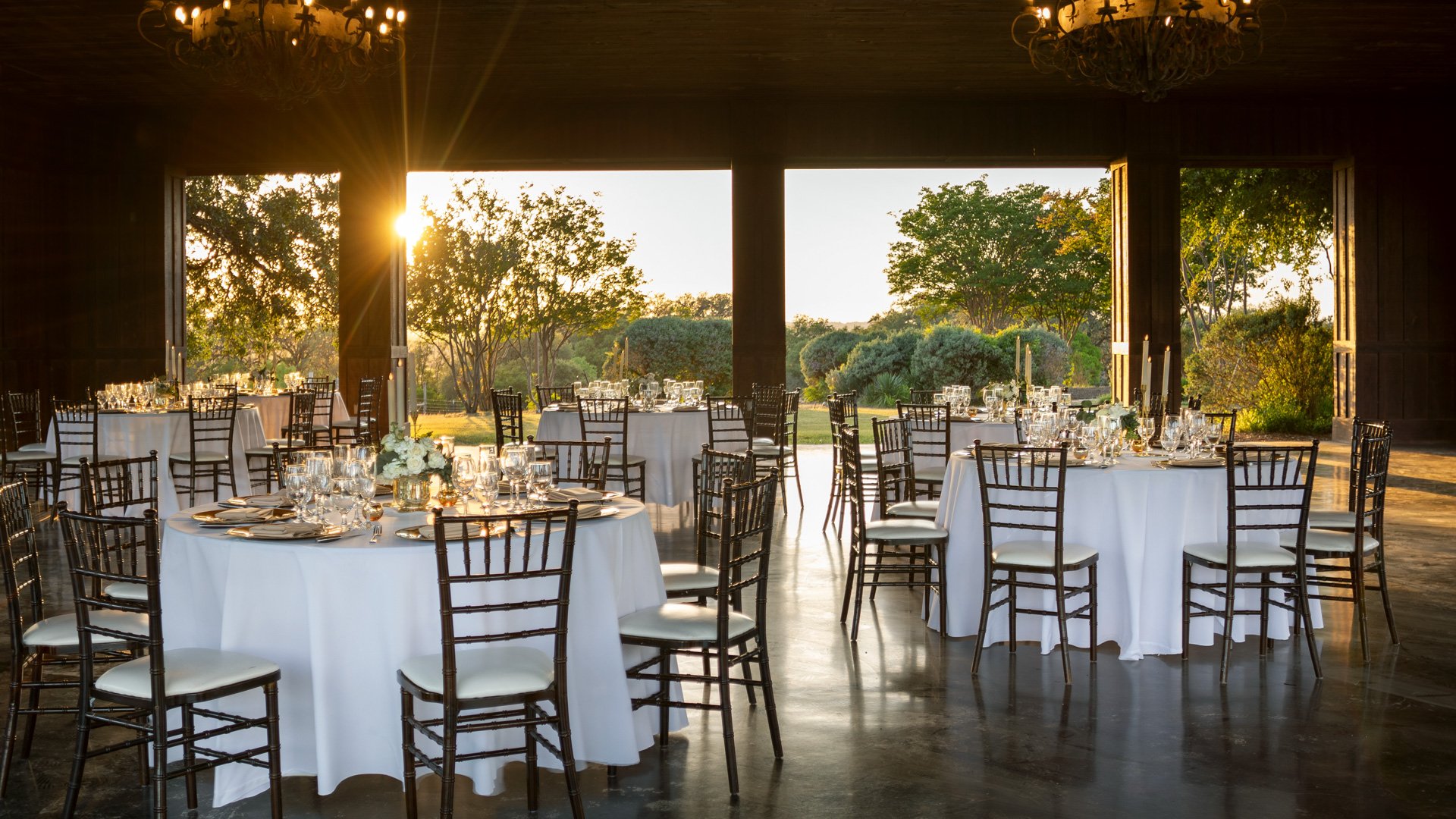 Romantic wedding reception at Hofmann Ranch’s Meadow Pavilion, featuring open-air views of the sunset, round tables with white linens, and rustic chandeliers overhead.