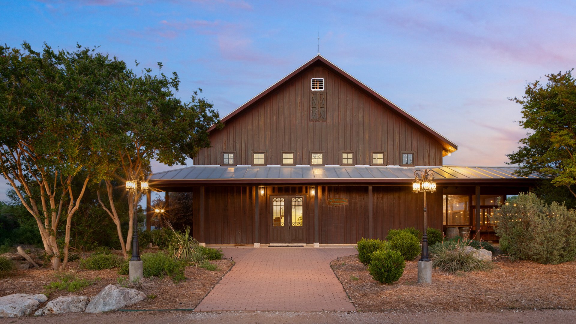 Evening view of Hofmann Ranch in Castroville, TX, showcasing its rustic barn-style exterior, warm lighting, and landscaped entrance surrounded by Texas greenery.