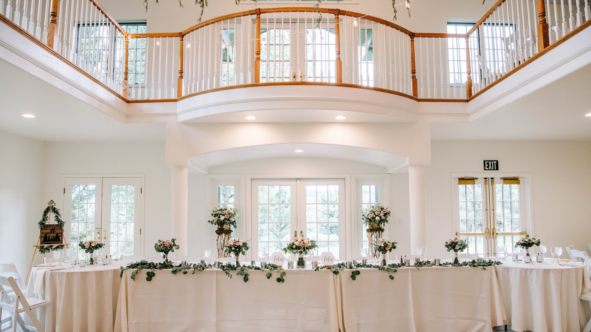 Elegant head table with cream linens and pink floral arrangements in bright two-story reception hall with balcony railings and natural light at Lionscrest Manor wedding venue