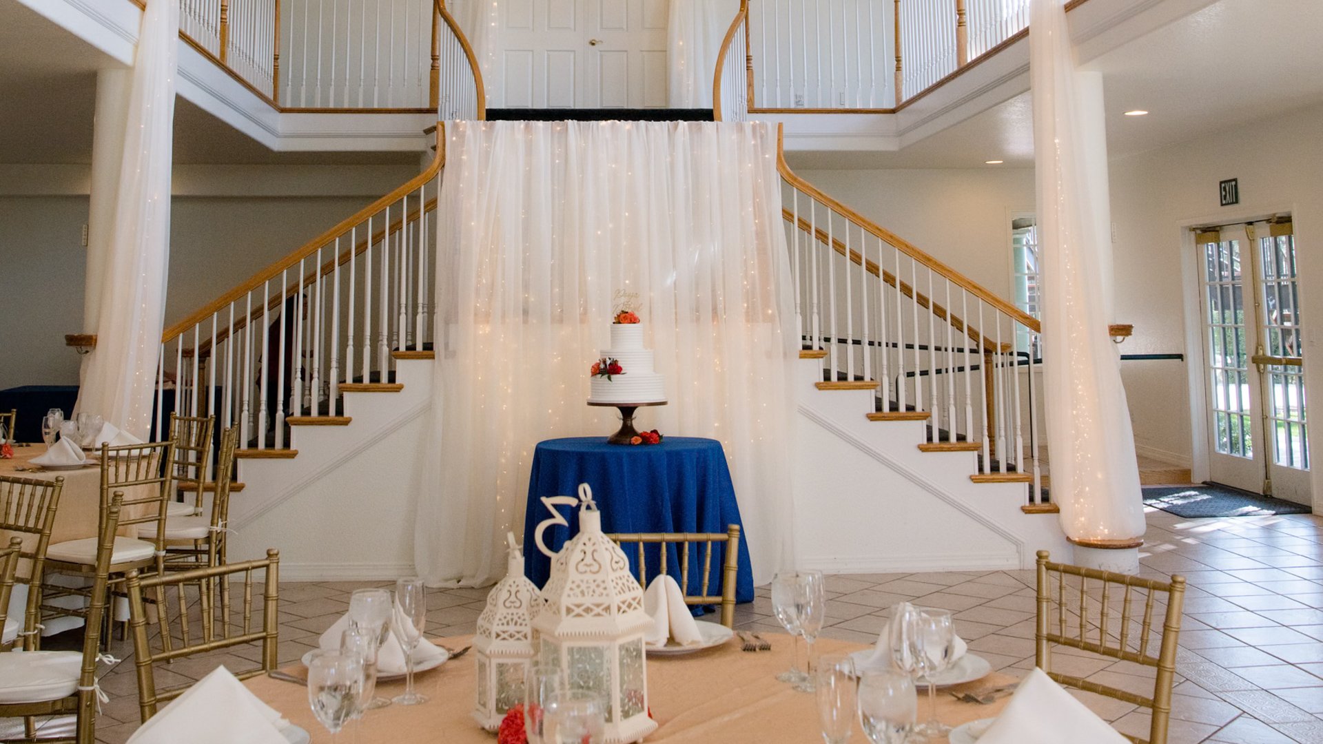 Elegant white wedding cake on navy blue tablecloth positioned between symmetrical staircases with white curtain backdrop and string lights, white lantern centerpieces on guest tables at Lionscrest Manor