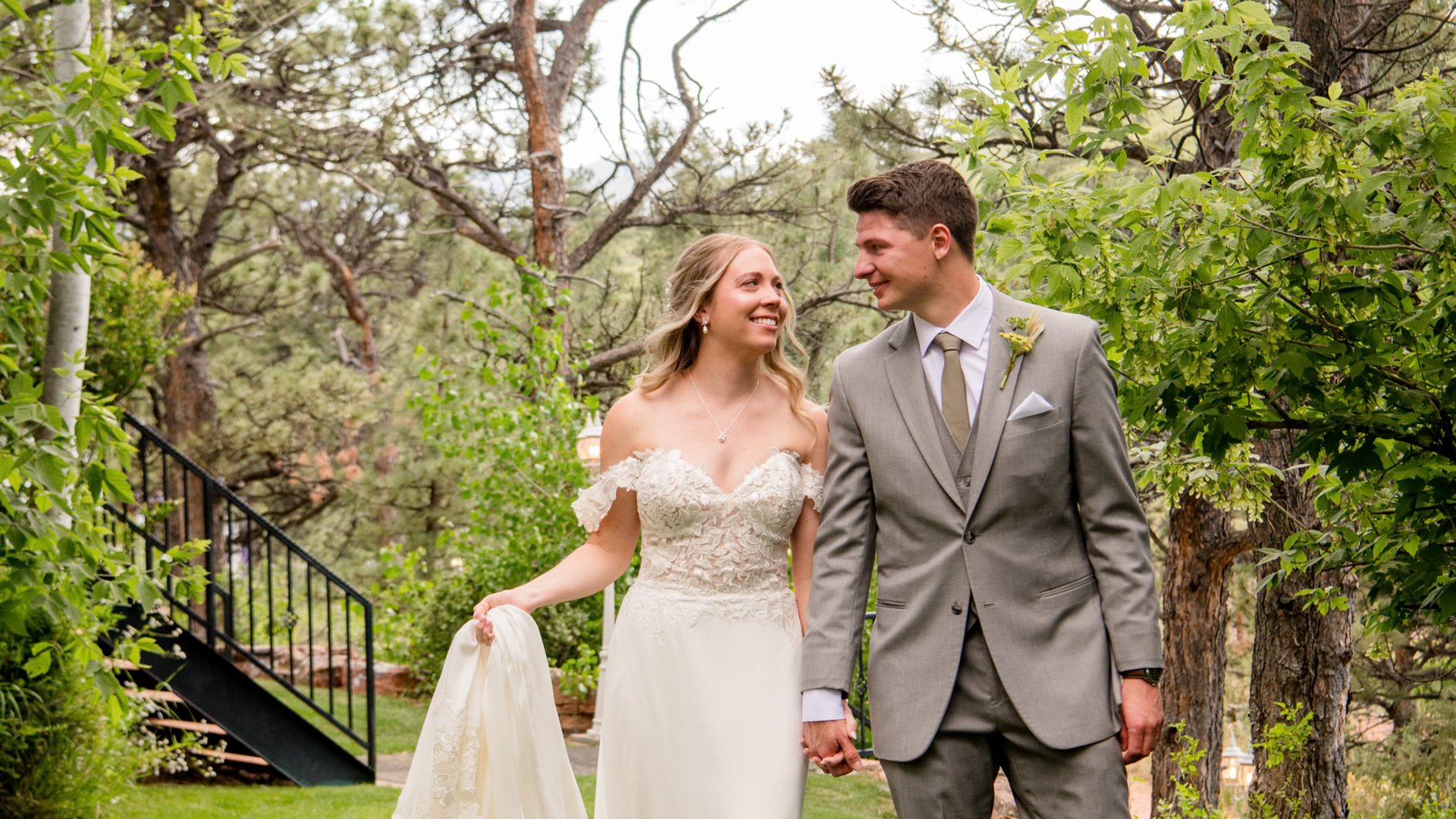 Smiling bride in off-shoulder lace gown and groom in grey suit with boutonniere walking hand in hand through lush forest garden with aspen and pine trees at Lionscrest Manor wedding venue in Lyons, Colorado