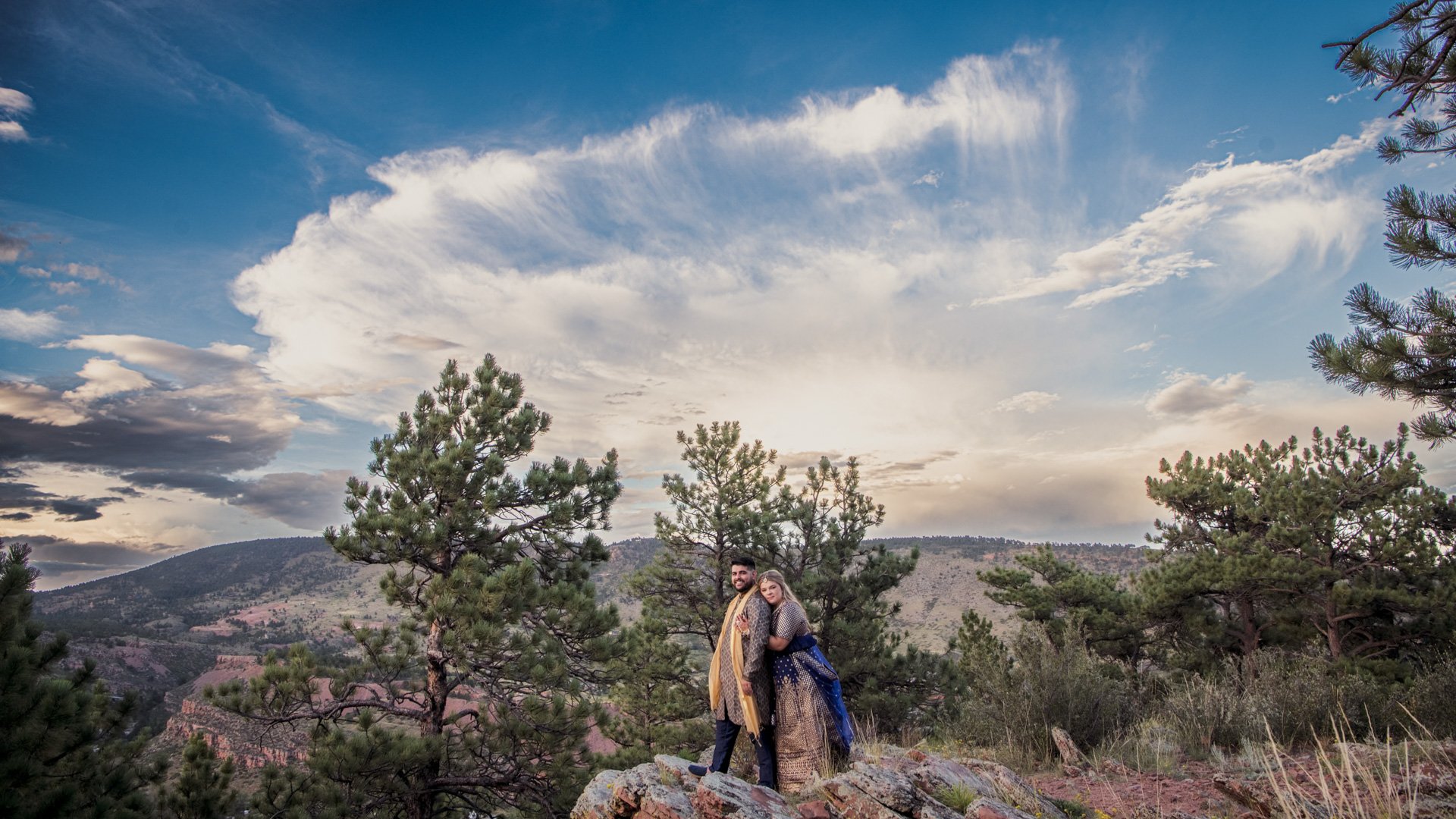 Couple in traditional Indian wedding attire embracing on rocky overlook with sweeping Colorado mountain valley views and dramatic cloud formations near Lionscrest Manor in Lyons