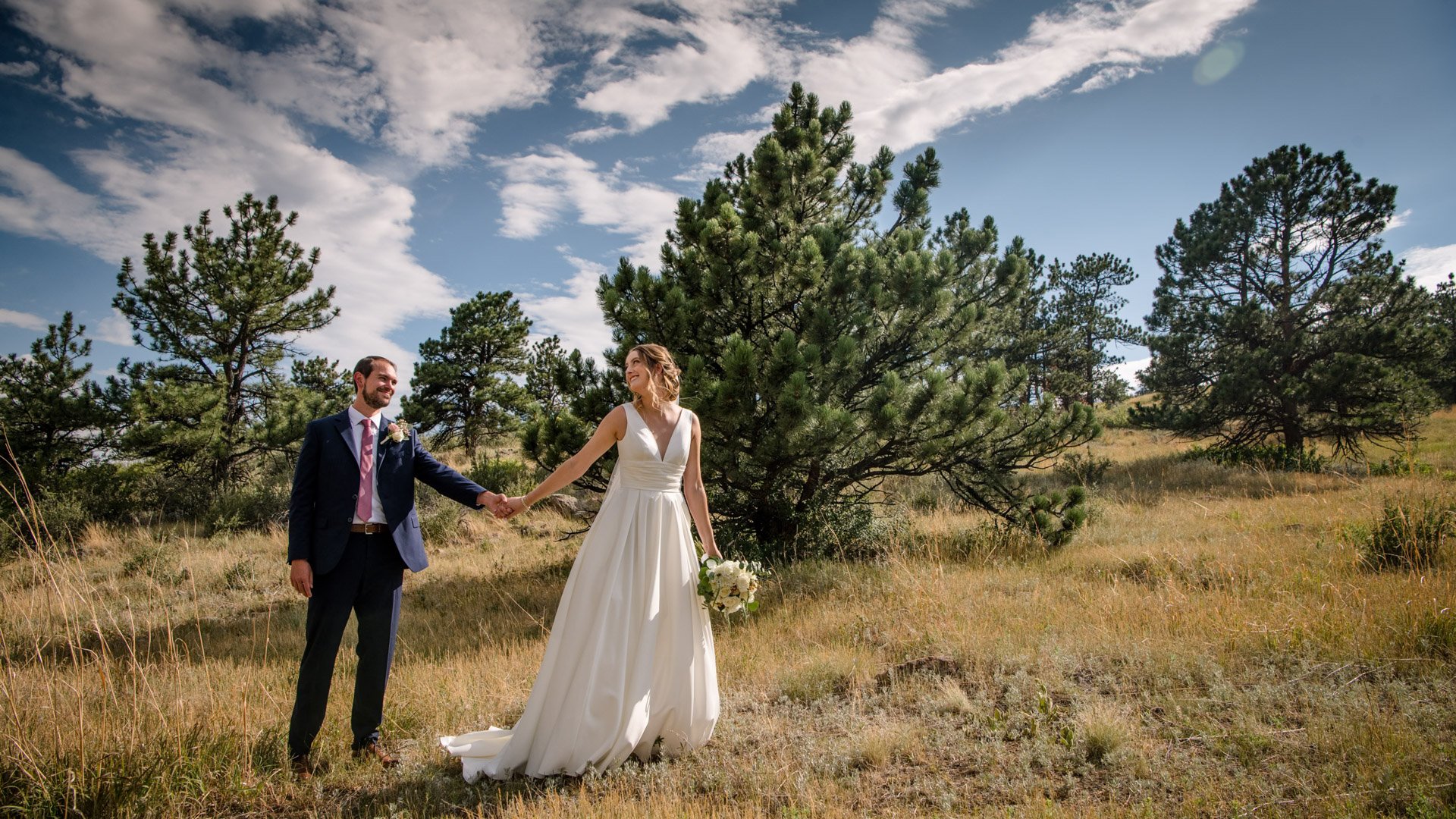 Bride in flowing white v-neck gown and groom in navy suit walking hand in hand through golden grass meadow with pine trees and dramatic blue sky near Lionscrest Manor in Colorado
