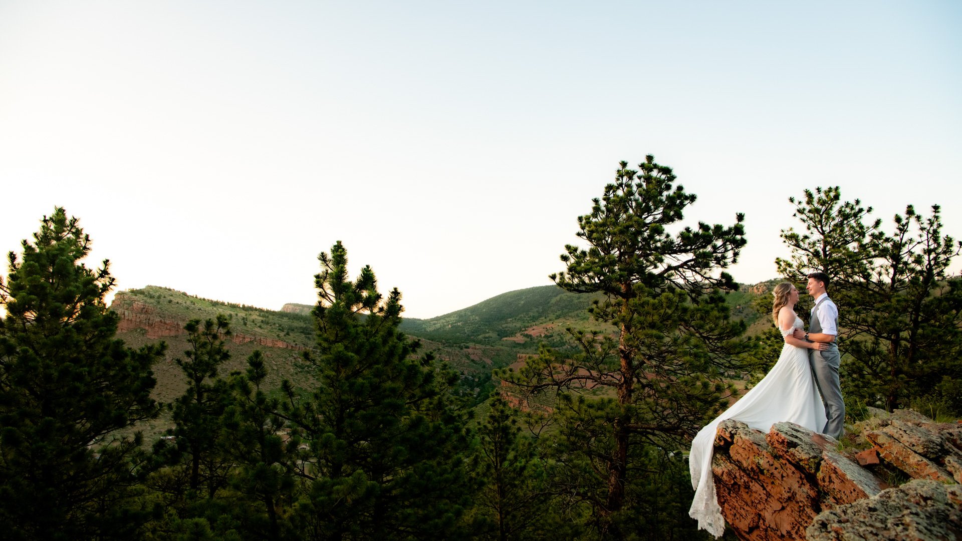 Bride in flowing white gown with dramatic train cascading over red rock cliff and groom in grey vest embracing on scenic overlook with sweeping Colorado mountain and pine forest views near Lionscrest Manor in Lyons
