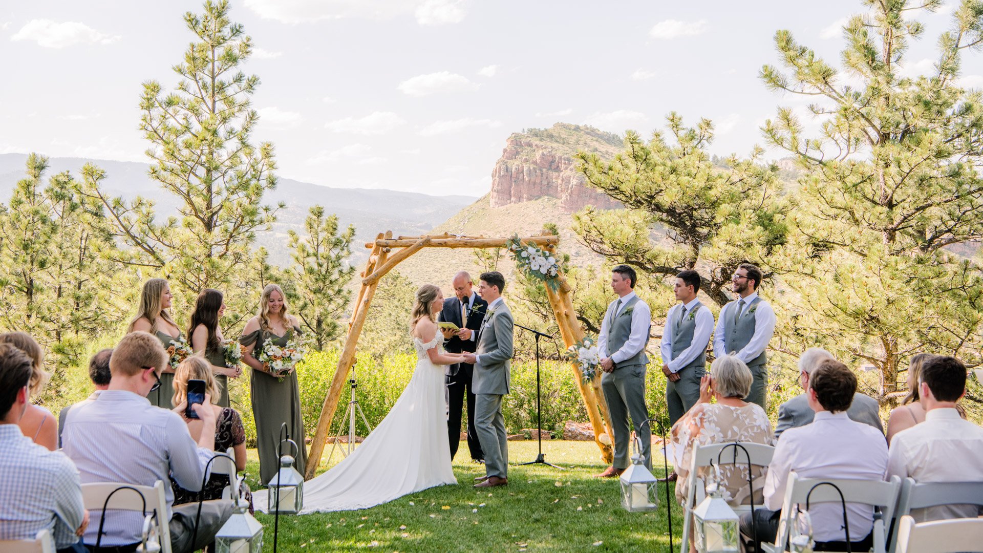Outdoor wedding ceremony with wooden arbor decorated with eucalyptus, bride and groom exchanging vows, wedding party and guests, iconic red rock mesa backdrop at Lionscrest Manor in Colorado