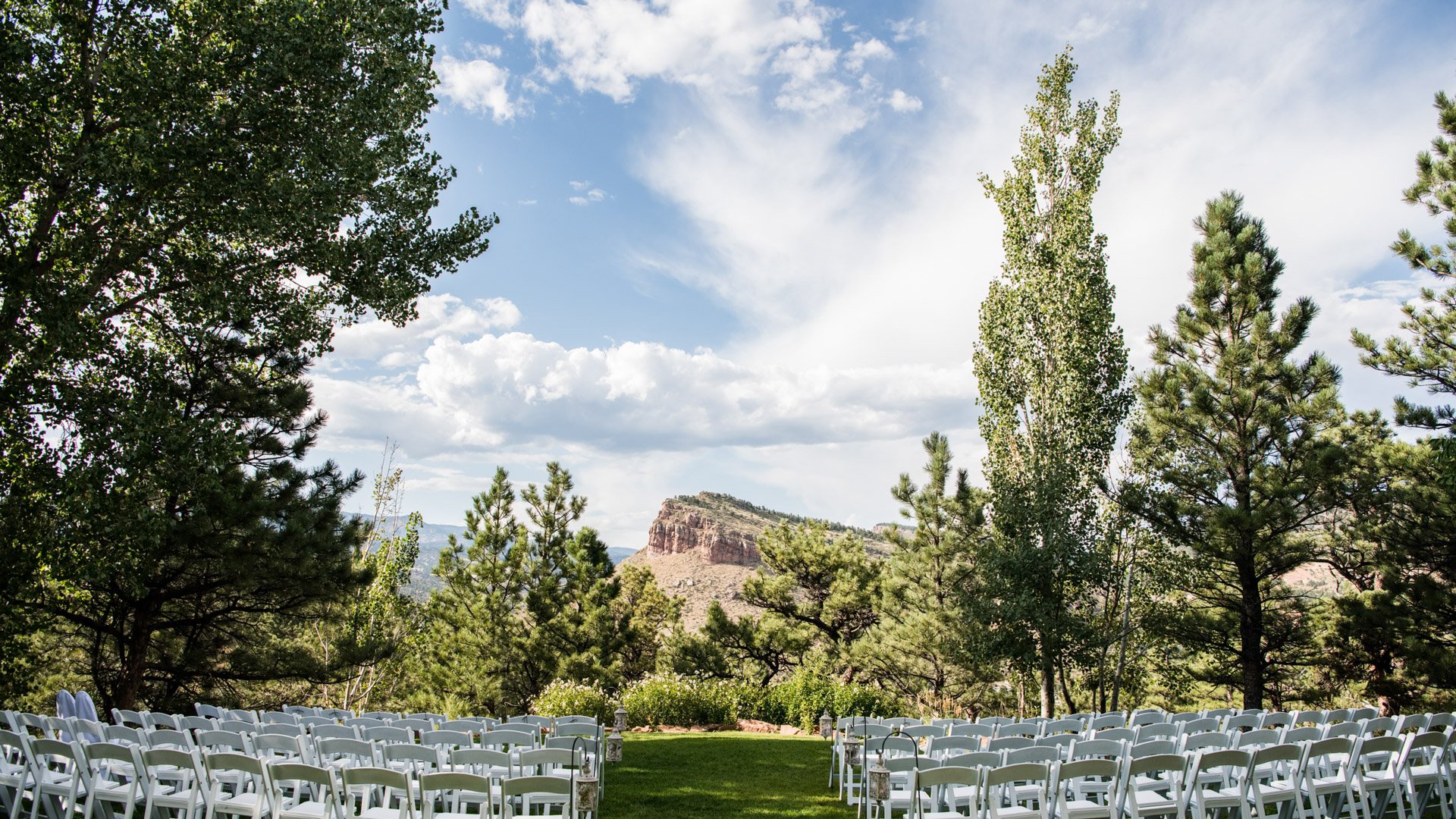 Outdoor wedding ceremony with white chairs arranged on green lawn overlooking dramatic Colorado mountain formations framed by pine and aspen trees at Lionscrest Manor in Lyons