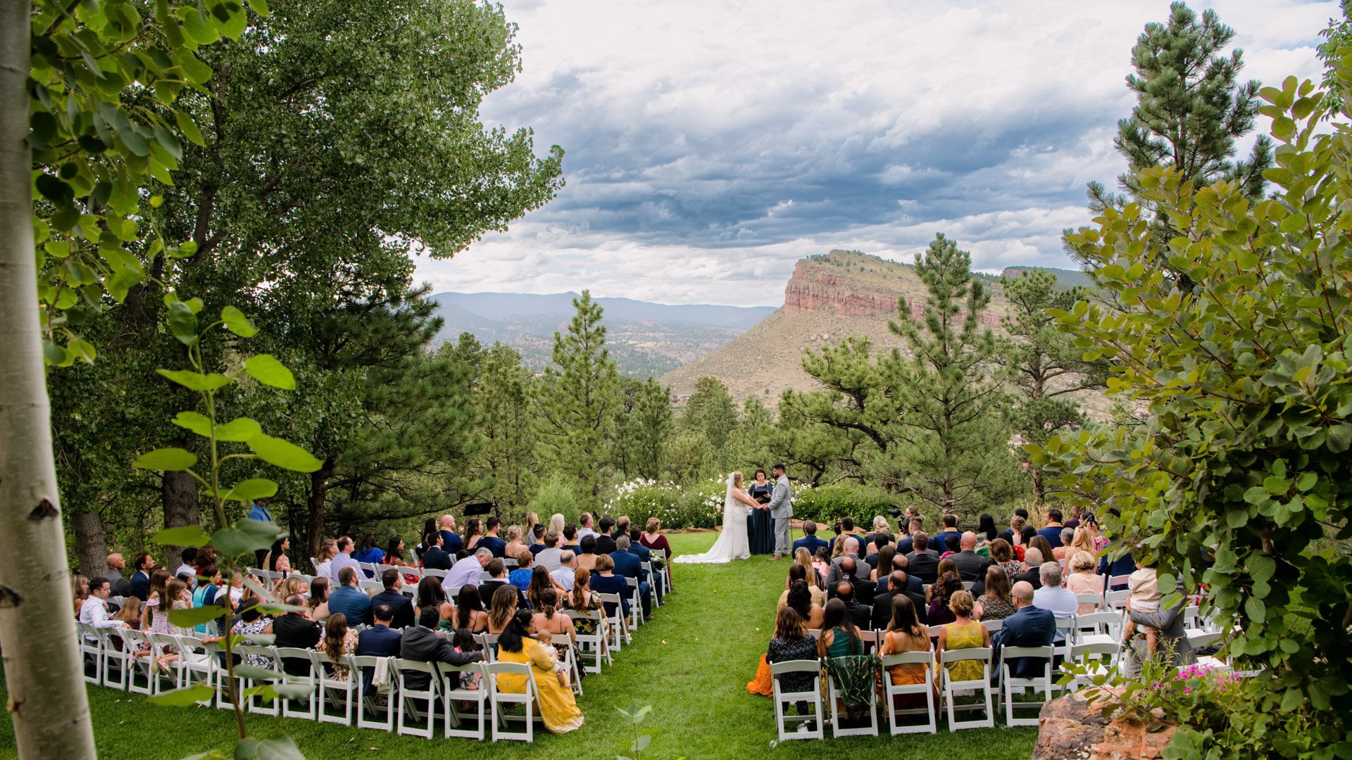 Outdoor wedding ceremony underway with guests seated in white chairs watching bride and groom exchange vows with dramatic Colorado mesa and mountain landscape framed by aspen and pine trees at Lionscrest Manor in Lyons