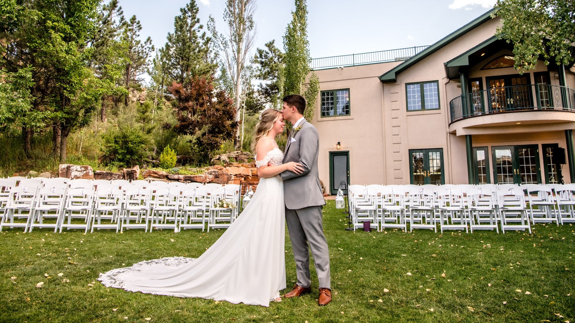 Bride with flowing cathedral train and groom in grey suit kissing on lawn in front of Lionscrest Manor venue building with ceremony chairs and natural rock garden landscaping visible