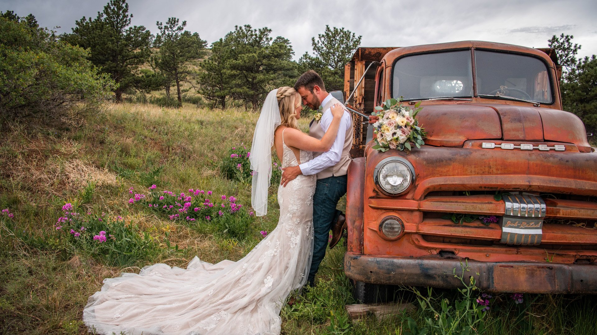 Bride in lace gown with cathedral train and groom embracing by weathered orange vintage Chevrolet truck with wildflowers and Colorado foothills at Lionscrest Manor wedding venue
