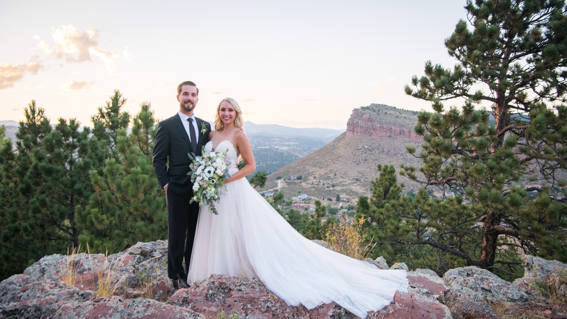 Bride in flowing white gown with cathedral train and groom in black suit posing on rocky overlook with dramatic Colorado mesa backdrop and pine trees at sunset near Lionscrest Manor