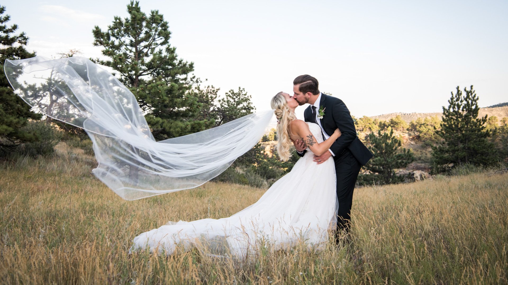 Bride in white ballgown with dramatically windswept cathedral veil and groom in black suit kissing in golden grass meadow with pine trees and Colorado foothills near Lionscrest Manor in Lyons