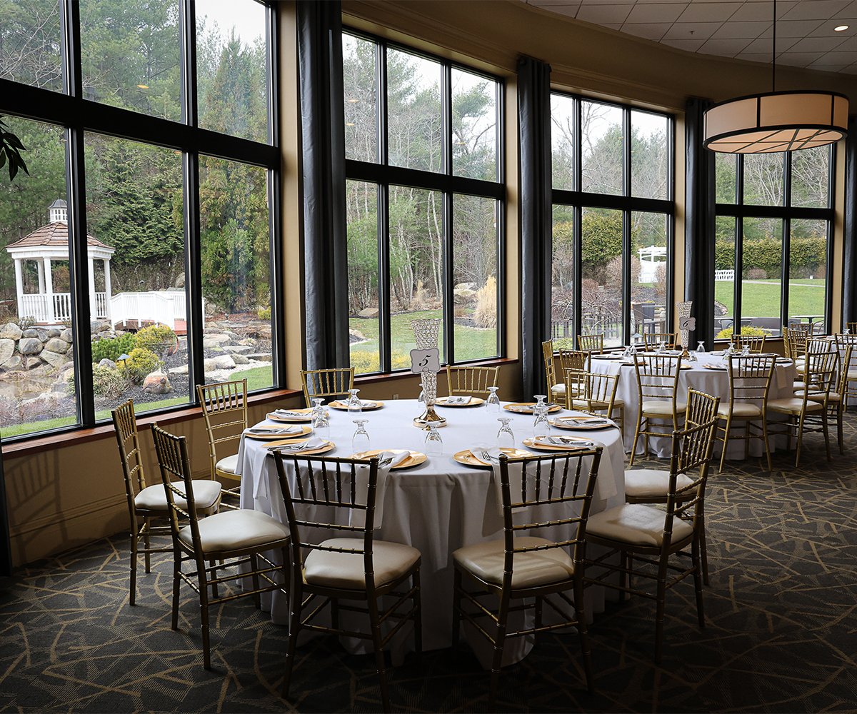 interior event space at Miraval Gardens by Wedgewood Weddings, with round tables, gold chiavari chairs, stone fireplace, chandeliers, and large windows providing natural light and views of the outdoor landscape