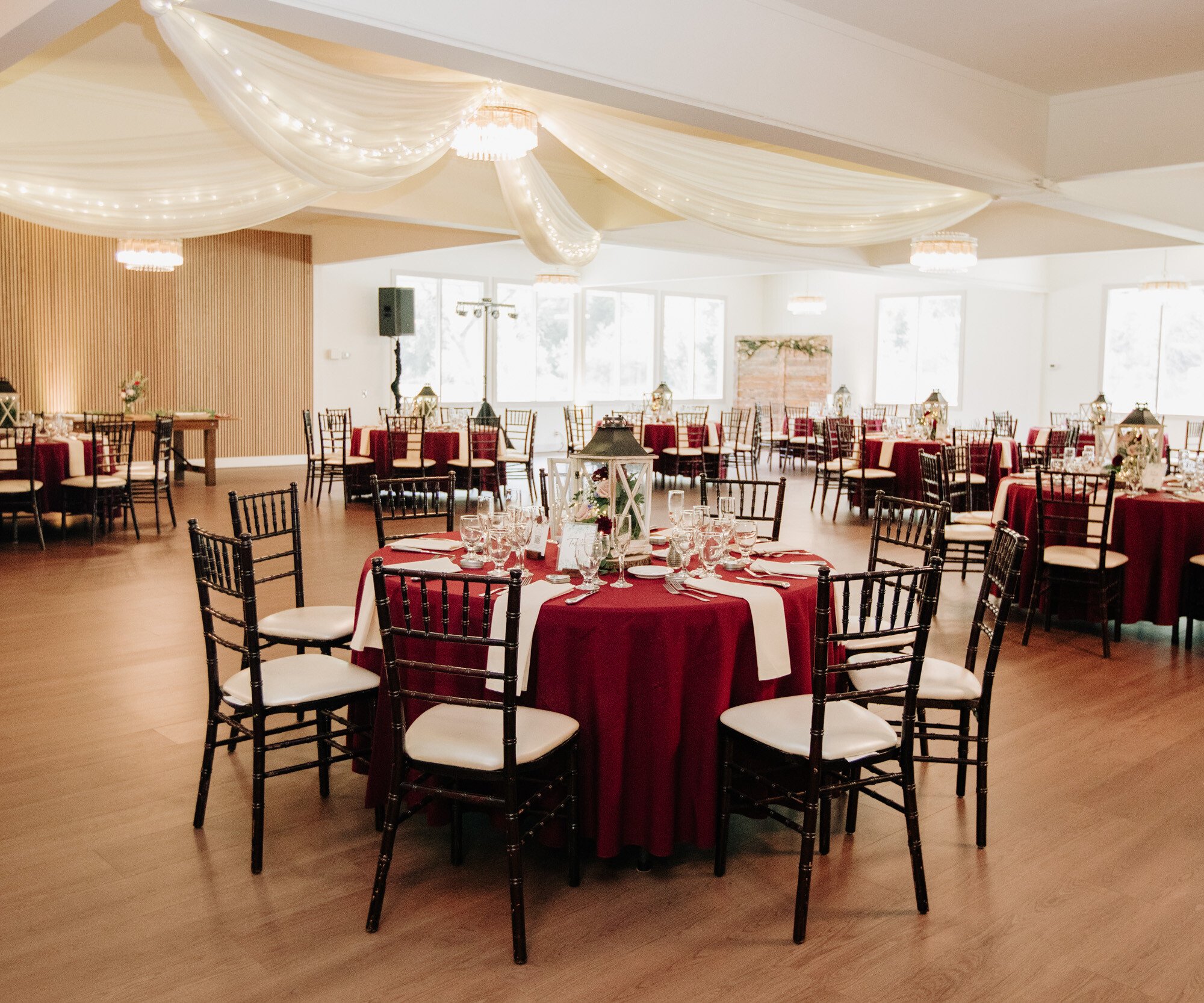 A spacious event hall with natural light streaming through large windows. Multiple round tables are dressed in burgundy tablecloths with white runners and surrounded by dark Chiavari chairs. White fabric drapes with string lights decorate the ceiling, creating an elegant atmosphere.
