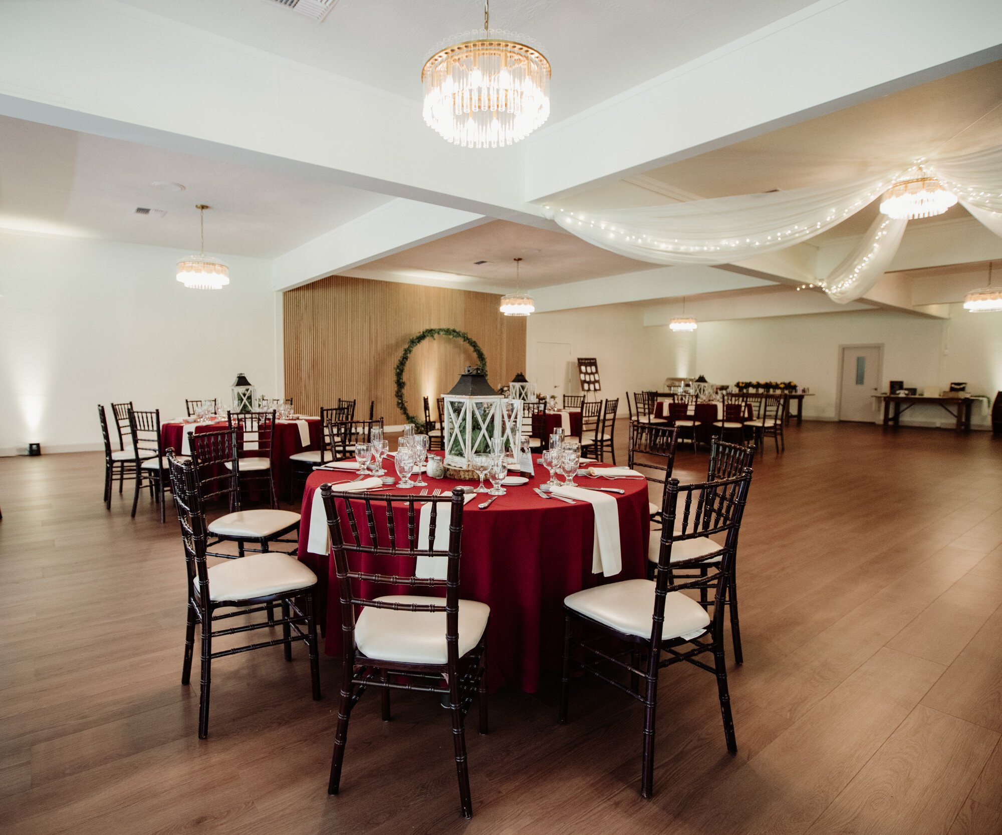 An elegant event space is set up for a formal dinner. Round tables are covered in burgundy tablecloths and white runners. The room features crystal chandeliers, draped white fabric with fairy lights on the ceiling, and a wooden accent wall with a round greenery arch.