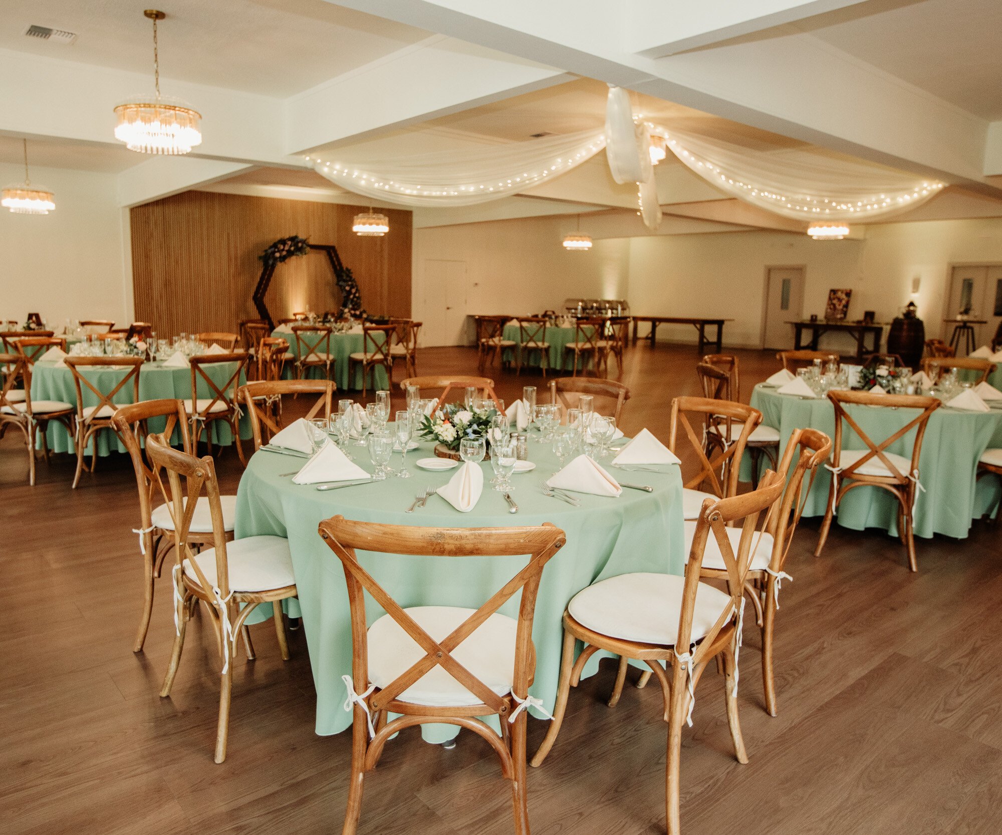 A wedding reception venue featuring round tables with mint green tablecloths and wooden cross-back chairs with white cushions. The ceiling is decorated with flowing white fabric and string lights. A hexagonal wooden arch with greenery serves as a backdrop feature.