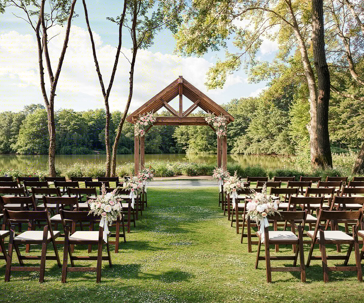 An outdoor ceremony space with a wooden arch decorated with flowers, positioned by a lake with rows of chairs creating an aisle.