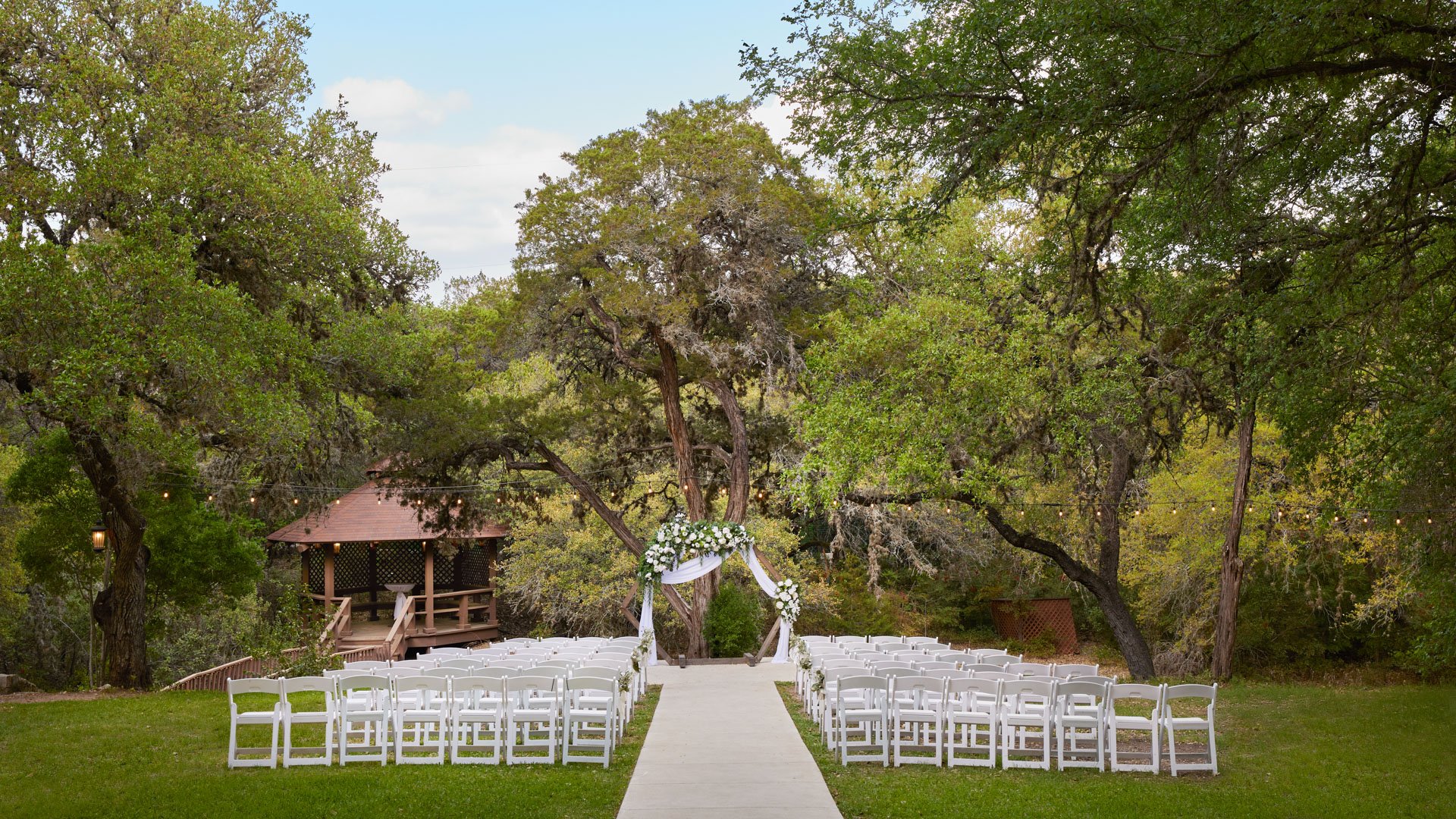Scenic Springs forest lawn ceremony space in Helotes, Texas, with floral arch, gazebo, white seating, and overhead string lights.