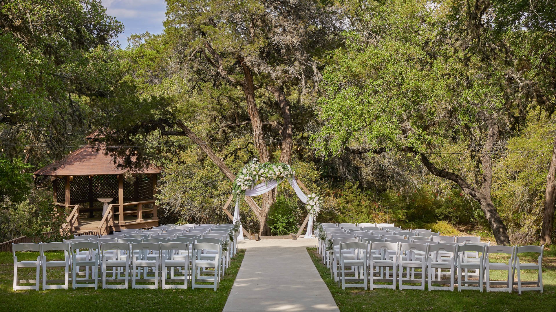 Outdoor ceremony space at Scenic Springs in Helotes, Texas, with floral hexagon arch, white chairs, and oak trees.