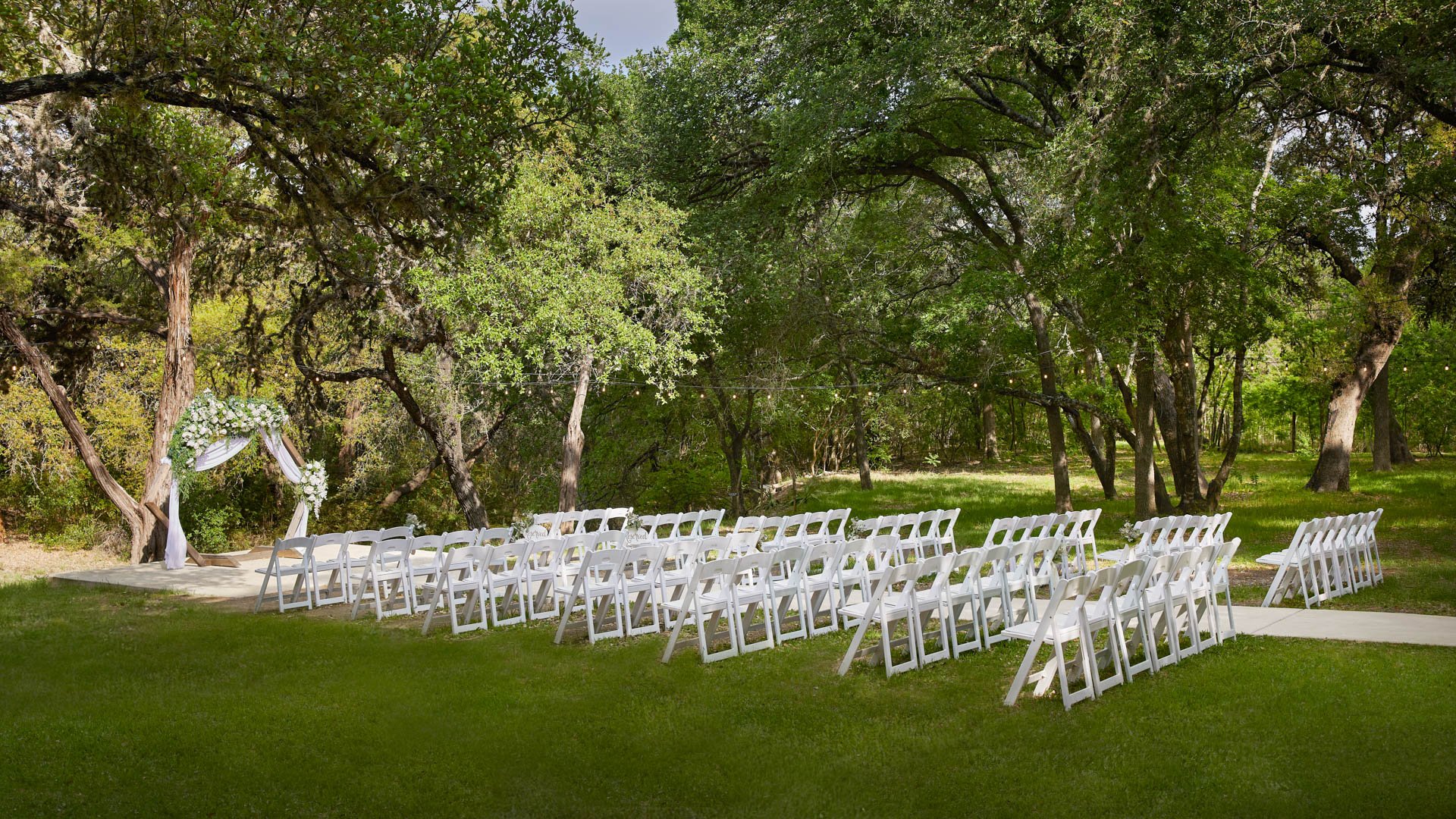 Side view of the Scenic Springs outdoor ceremony site in Helotes, Texas, featuring a floral arch and rows of white chairs surrounded by trees.