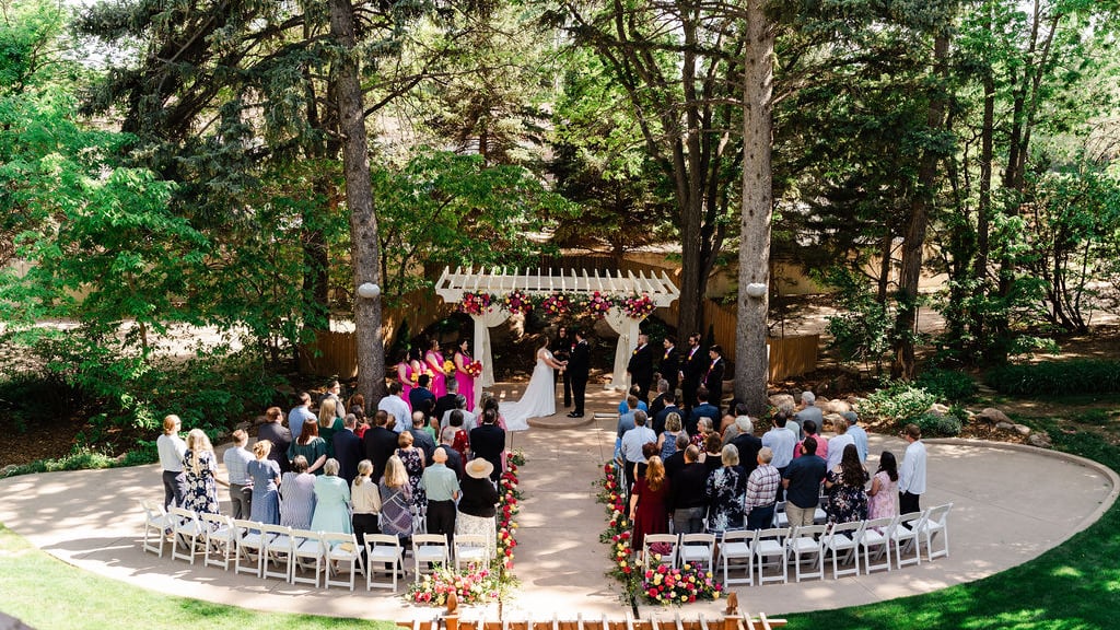 Outdoor summer ceremony at Tapestry House by Wedgewood Weddings in Northern Colorado. Featuring red and white flowers and a white pergola surrounded by mature trees.