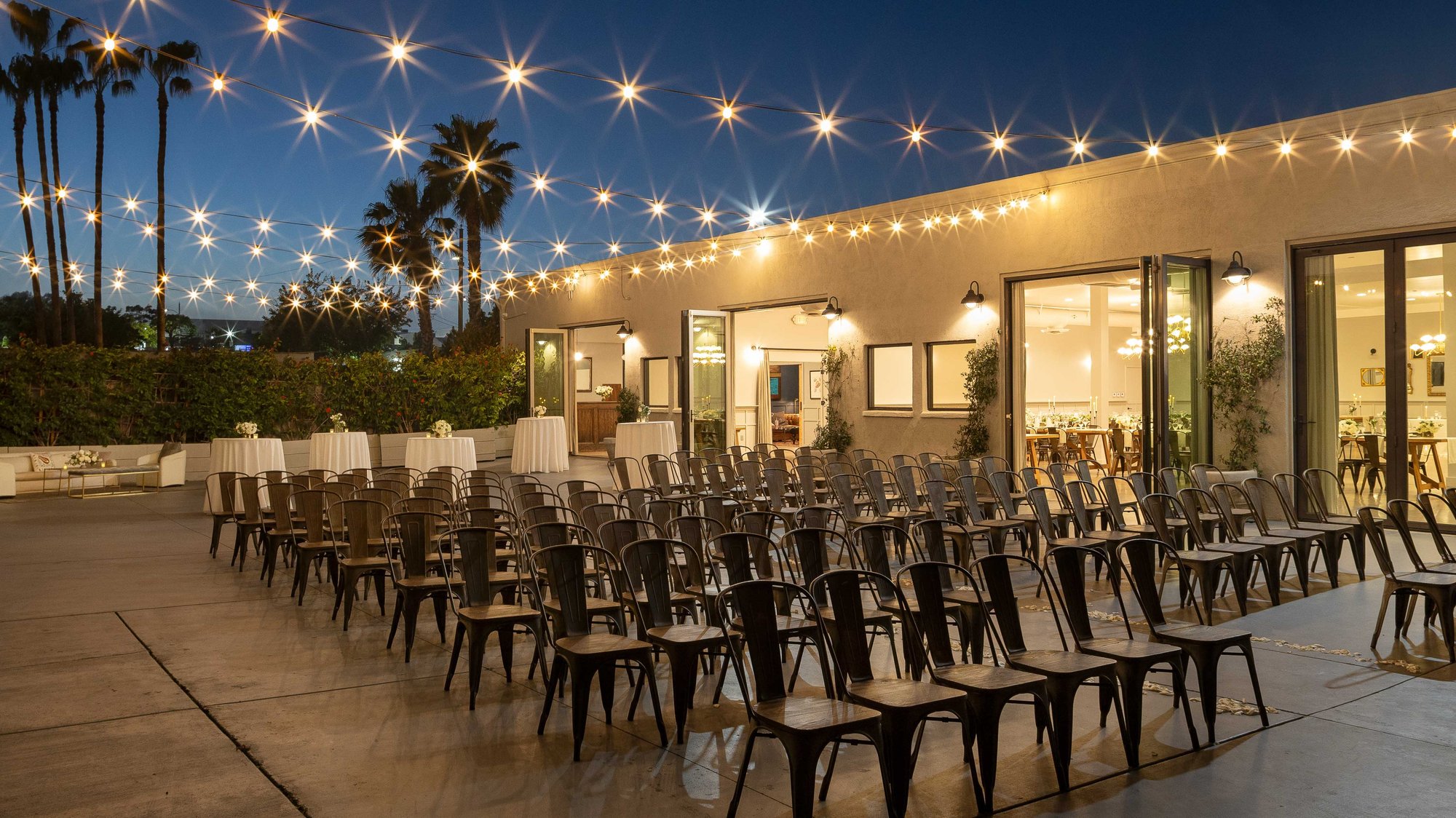 An evening outdoor ceremony setup with string lights, metal chairs, and palm trees creating a Southern California ambiance