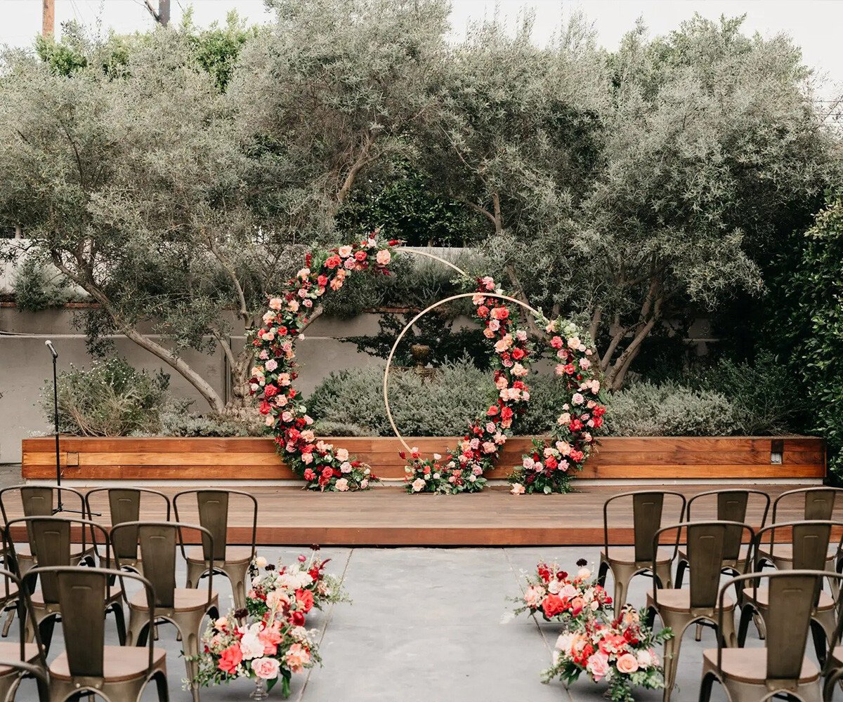 Modern ceremony at The Harper Orange County: circular floral arch with coral roses, industrial chairs, wood stage backdrop, outdoor elegance