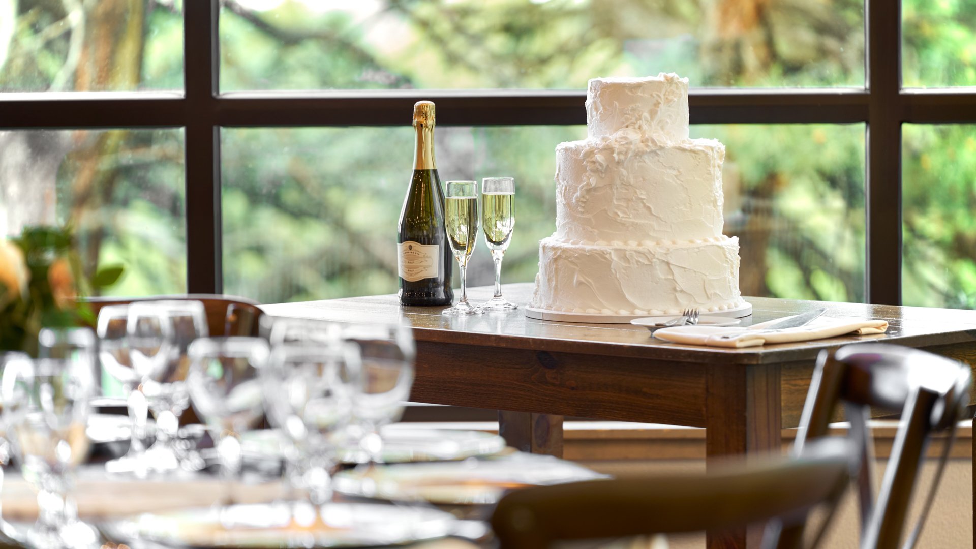 Three-tier white wedding cake with champagne setup on a wooden table at The Pines reception room overlooking lush green trees.