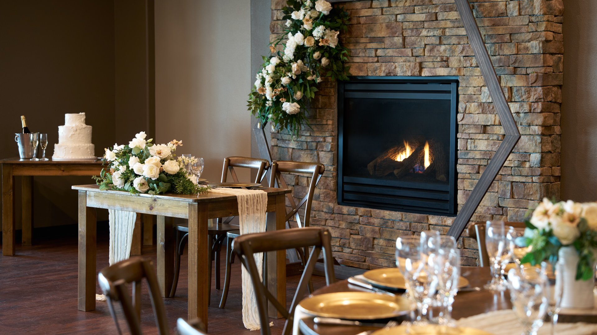 Warm and elegant wedding reception setup at The Pines in Colorado featuring a stone fireplace adorned with white floral arrangements, a sweetheart table, and a tiered wedding cake.