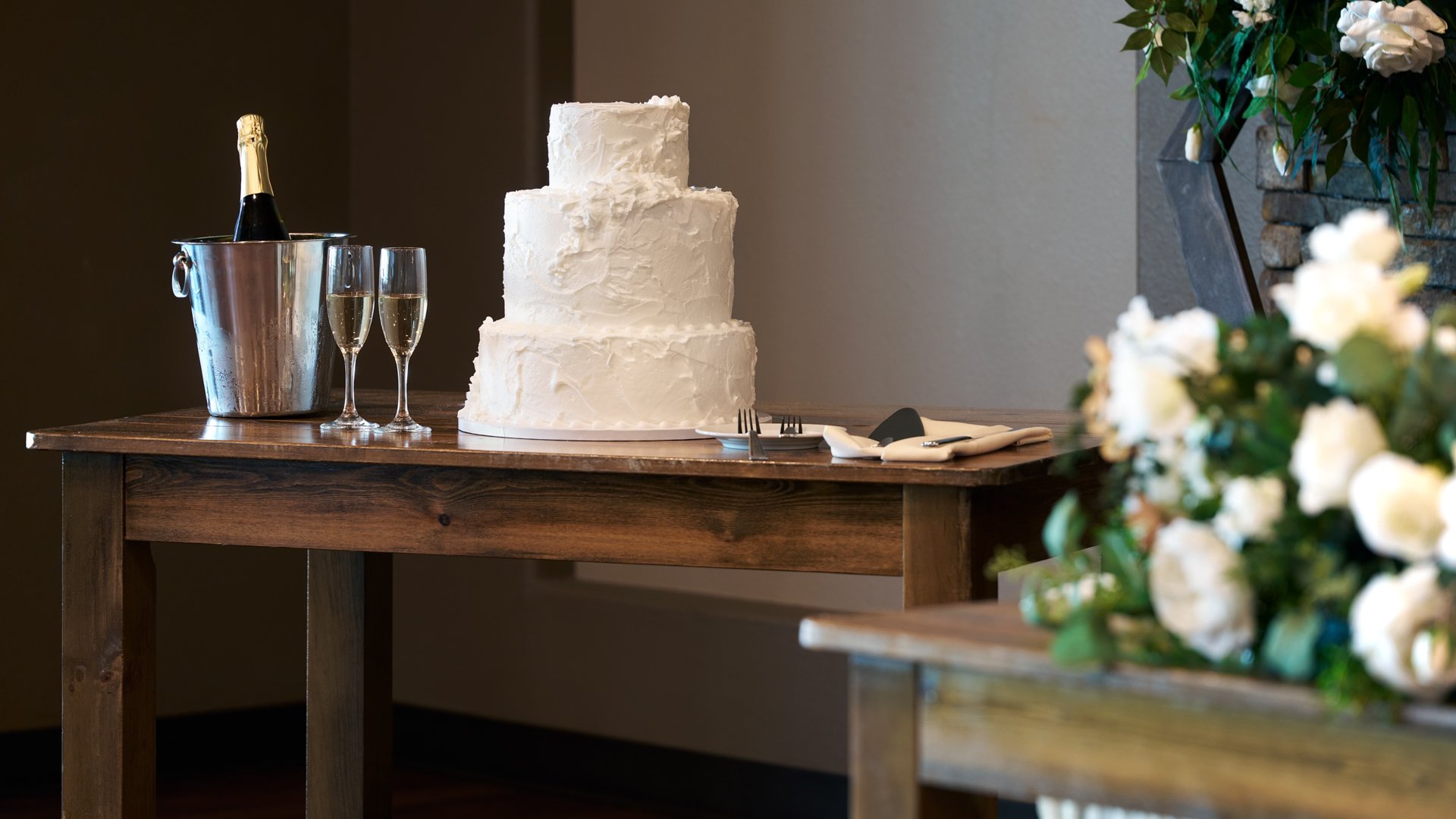 Close-up of a three-tier white wedding cake with champagne and flutes on a rustic wooden table at The Pines reception room.