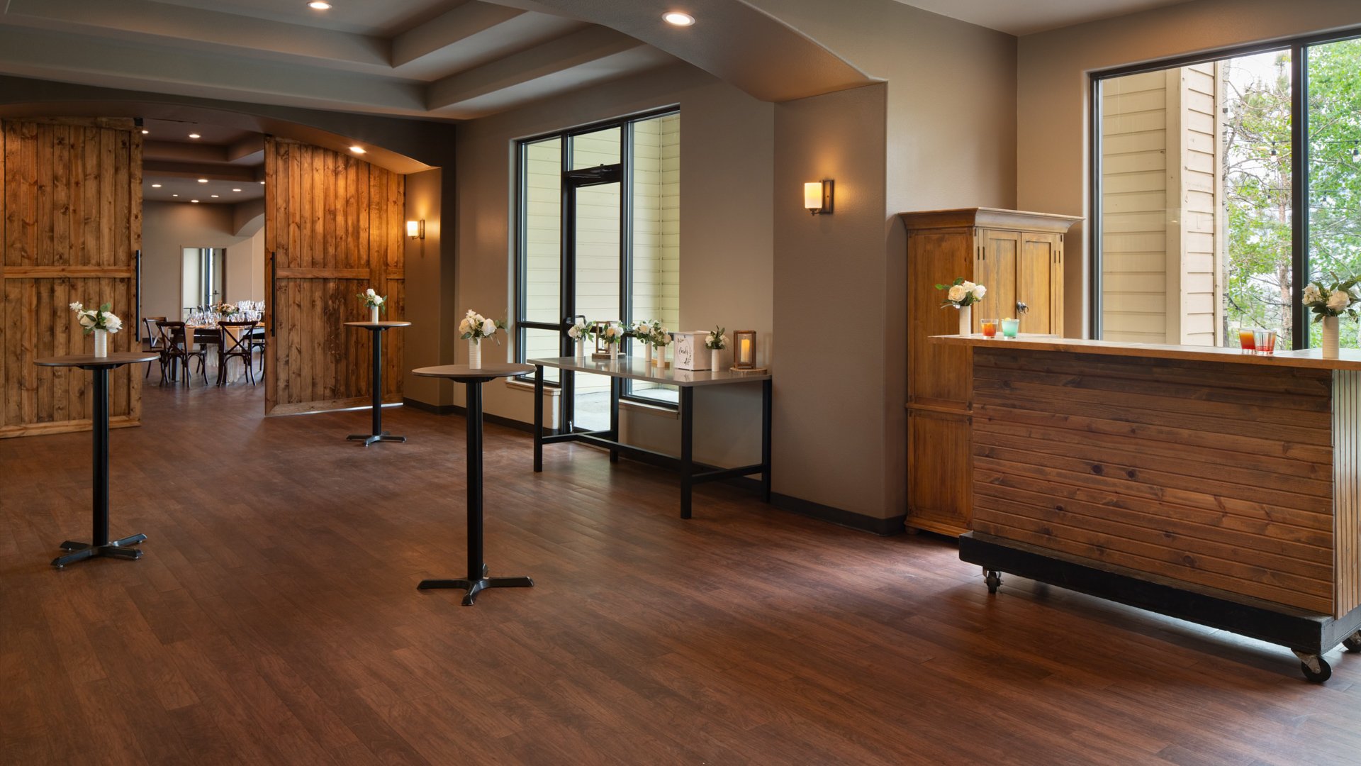 Elegant cocktail hour setup at The Pines featuring barn-style wooden doors, tall bistro tables with floral vases, and large windows overlooking the forest.