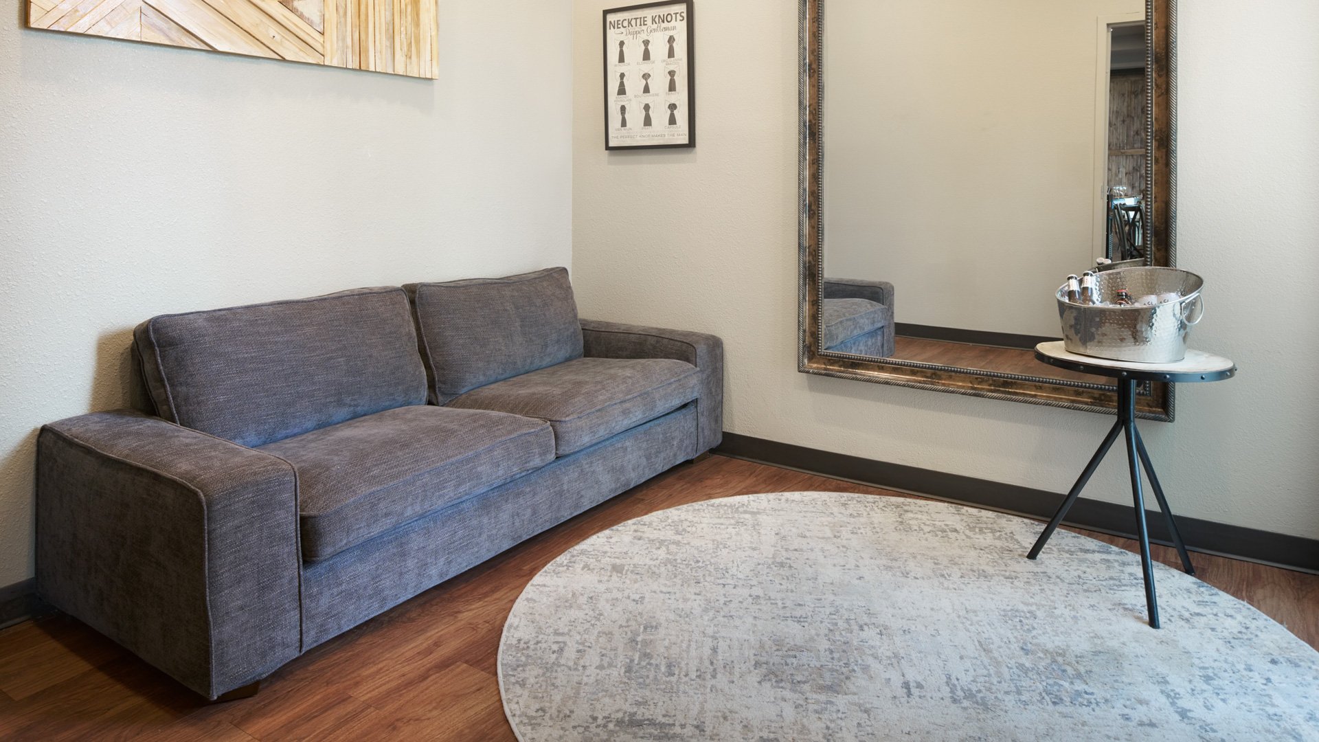 Groom’s lounge suite at The Pines featuring a gray couch, full-length mirror, modern art décor, and a metal beverage bucket on a round side table.