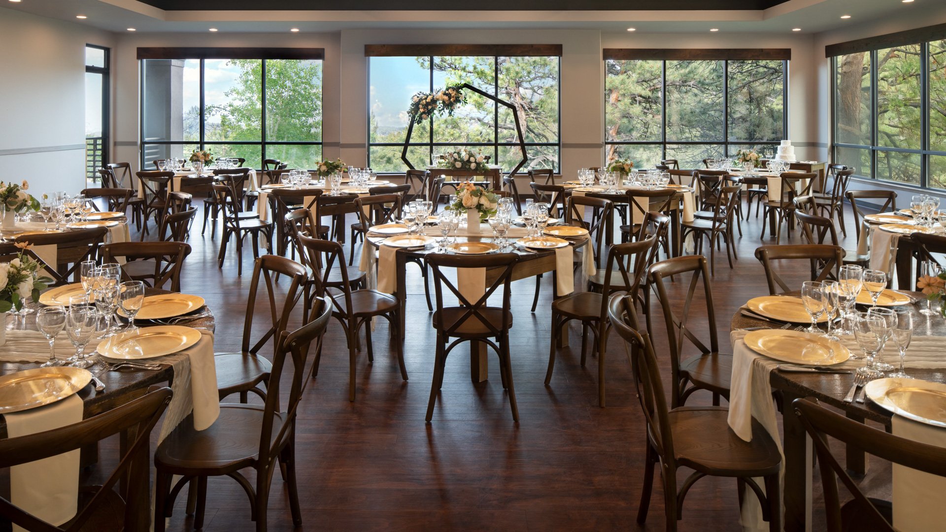Indoor wedding reception at The Pines Upper Level with floor-to-ceiling windows, wooden tables, gold chargers, and mountain forest views.