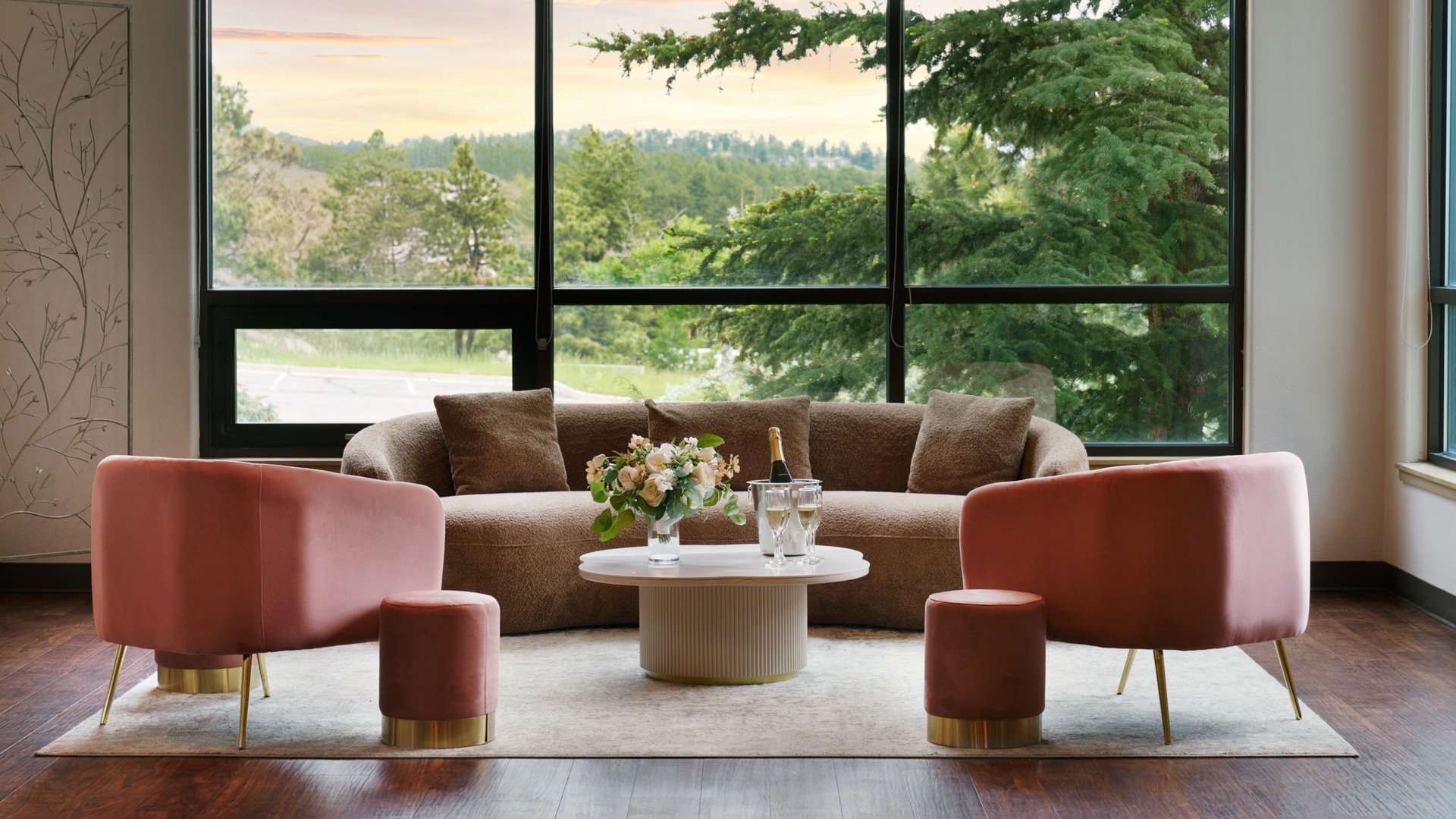 Sophisticated bridal suite with soft pink velvet chairs, a tan sofa, champagne setup, and large windows showcasing mountain and pine tree views.