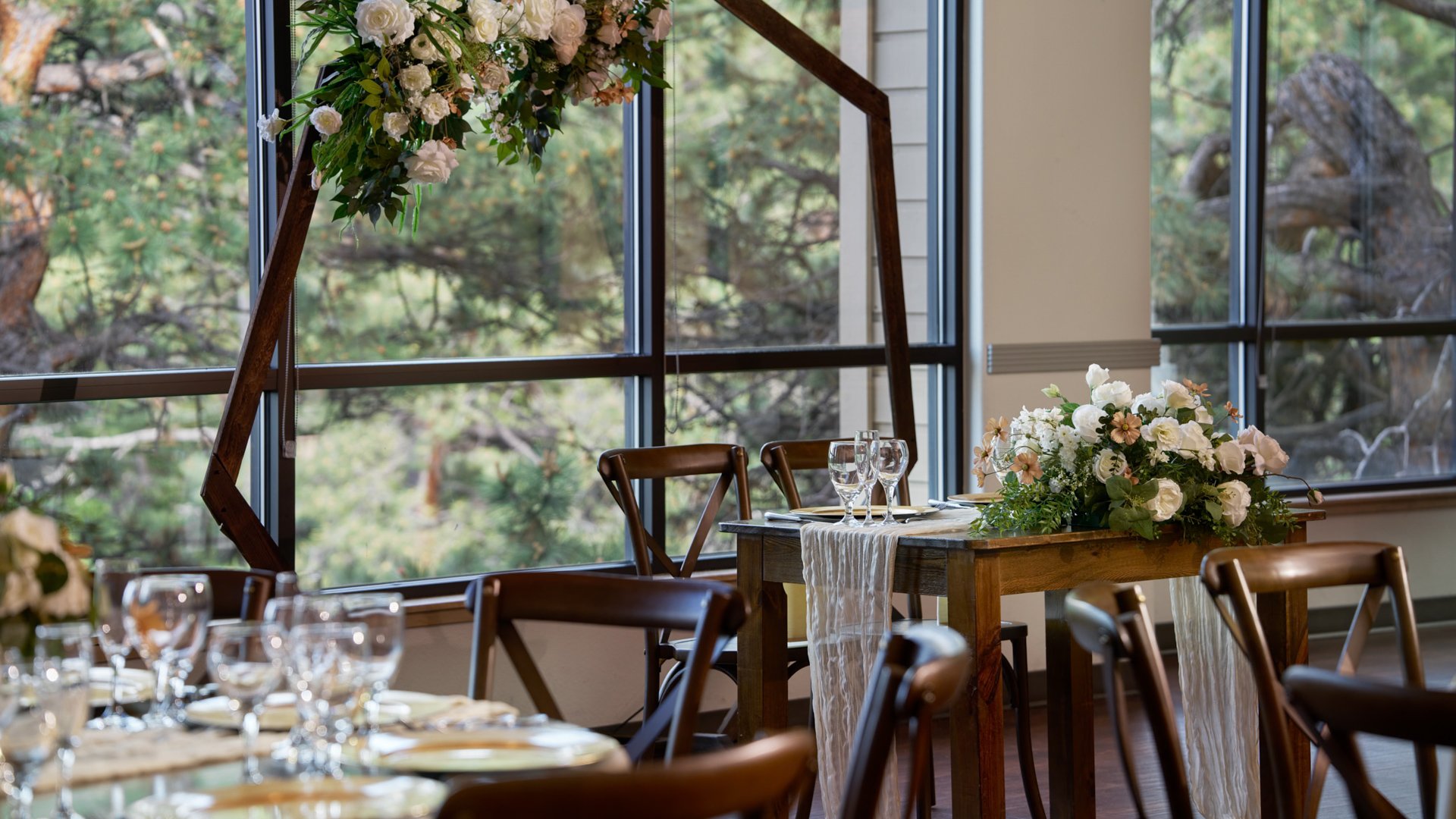 Sweetheart table at The Pines adorned with white and blush florals, rustic wooden chairs, and a geometric floral arch beside large windows overlooking forest scenery.